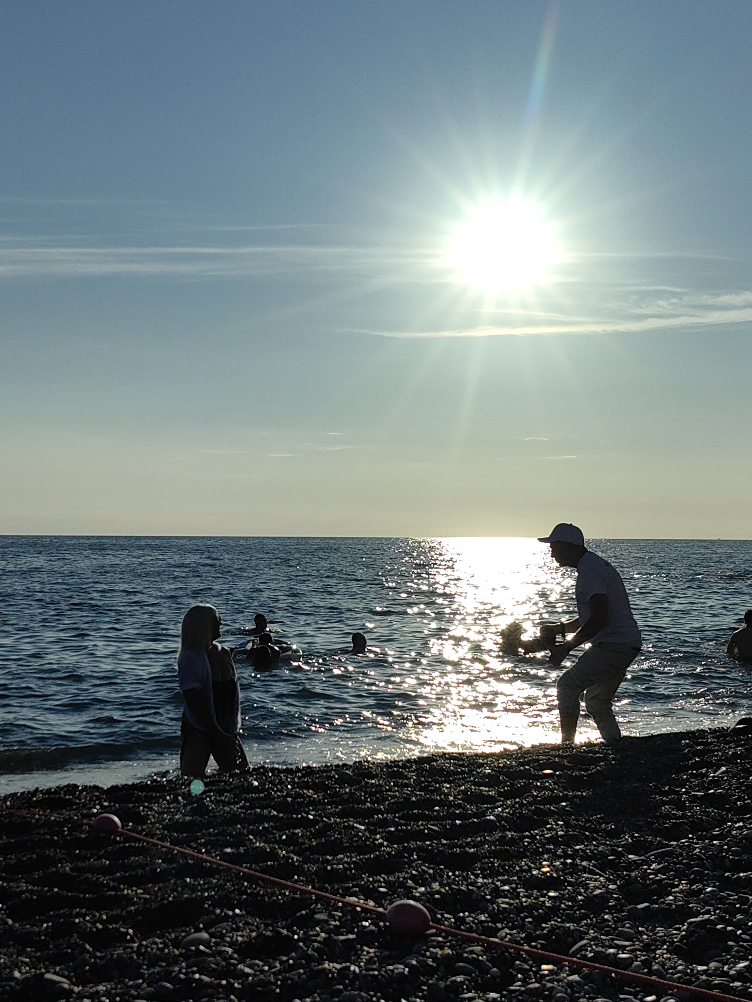 The image depicts a serene beach scene in Sochi, Russia, bathed in the warm glow of a setting sun. The sky is a clear, pale blue, with a few wispy clouds streaking across it. The sun, a brilliant starburst of light, dominates the upper right portion of the frame, casting shimmering reflections on the gentle waves of the Black Sea.
In the foreground, the pebbled beach is dark and rich in texture. A few red buoys are scattered along the shoreline, connected by thin ropes, possibly marking a swimming area or part of a fishing net.
On the beach, silhouetted against the bright sea, stand two figures. To the left, a person with blonde hair looks out towards the water, their posture relaxed. To the right, a person wearing a cap and t-shirt is captured in motion, seemingly holding a piece of equipment, perhaps a camera, and looking towards the sea.
In the water, several other figures are visible, enjoying the sea. Some are swimming or wading, while others are floating on inflatable rings. The water is a deep blue, with the sunlight creating a dazzling path of light across its surface.
The overall mood of the image is peaceful and idyllic, capturing a moment of leisure and enjoyment by the sea. The time of day is evident from the low angle of the sun and the warm, golden light. The clear sky and calm sea suggest pleasant weather.