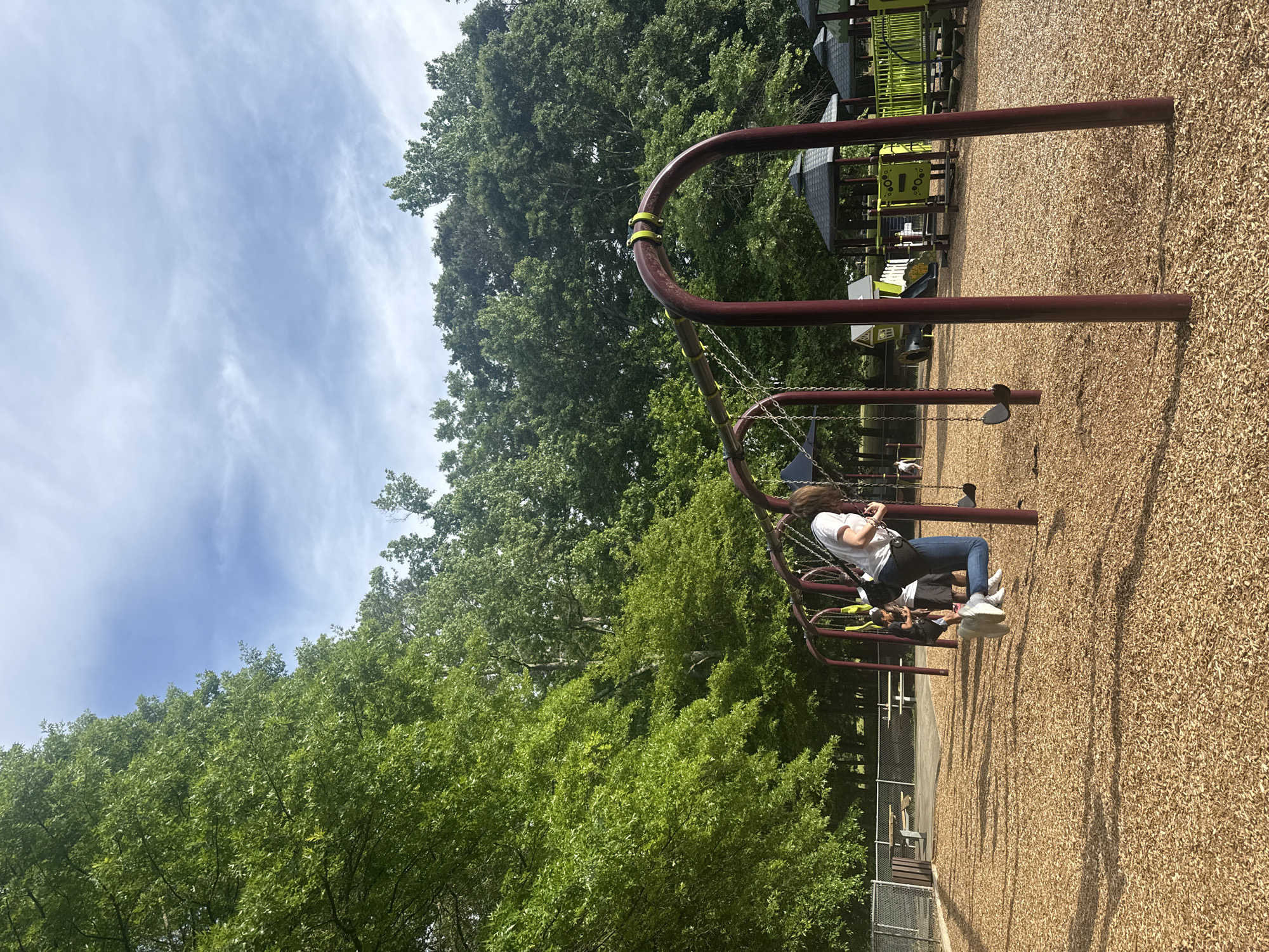 The image depicts a playground scene in Mountain Park, United States of America. Two individuals are on swings, with a person in a white t-shirt and blue jeans more prominently visible on a swing closer to the camera. The playground surface is covered in wood chips. In the background are various playground structures in shades of green and gray. Dense green trees surround the playground, and the sky has a mix of blue and scattered clouds. A fence is visible in the background.