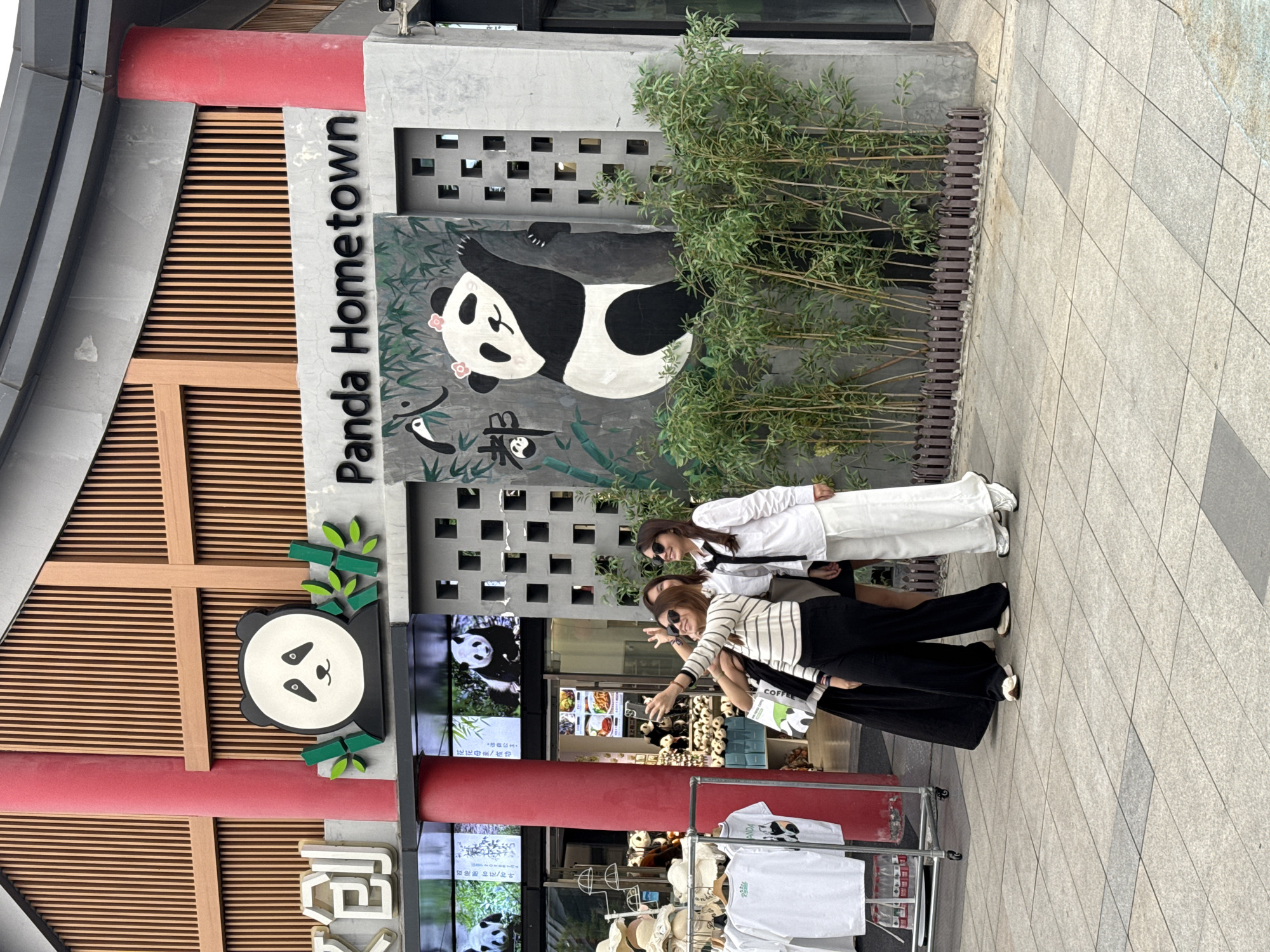 The scene is a commercial storefront identified as “Panda Hometown,” possibly a souvenir or theme shop, located in what appears to be Chenghua District, China, given the signage featuring pandas and Chinese characters. Three women are posing in front of the store, possibly taking photos. One woman is wearing a white shirt and white pants, while the other two wear striped shirts and black pants. A merchandise display with clothes, hats, and water bottles is situated in the foreground. The store’s facade includes panda-themed decorations, a painting of a reclining panda, and a green bamboo-like feature. A menu board or screen displaying panda images and food items is visible inside the store.