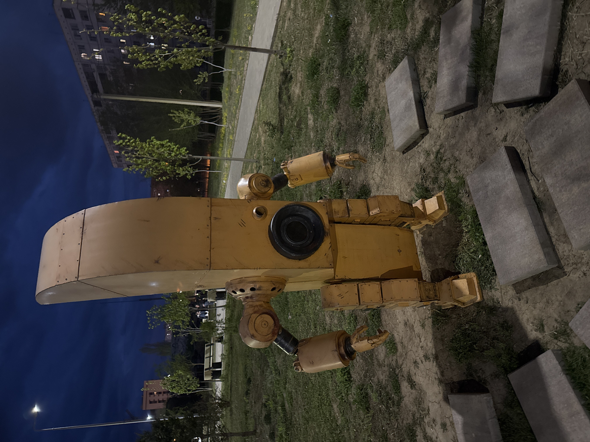 The image shows a yellow robot sculpture situated on grassy ground, likely an art installation. The robot is made of metal plates, with a rounded head, a cylindrical body, and articulated arms ending in claw-like hands. A large black circular aperture is visible on the robot's body. Several rectangular concrete slabs are arranged nearby, appearing as stepping stones. In the background, there is a sidewalk bordered by grass, and a building with illuminated windows stands against a dark blue sky. The location is likely the city of Volzhsky, Russia, based on the request.