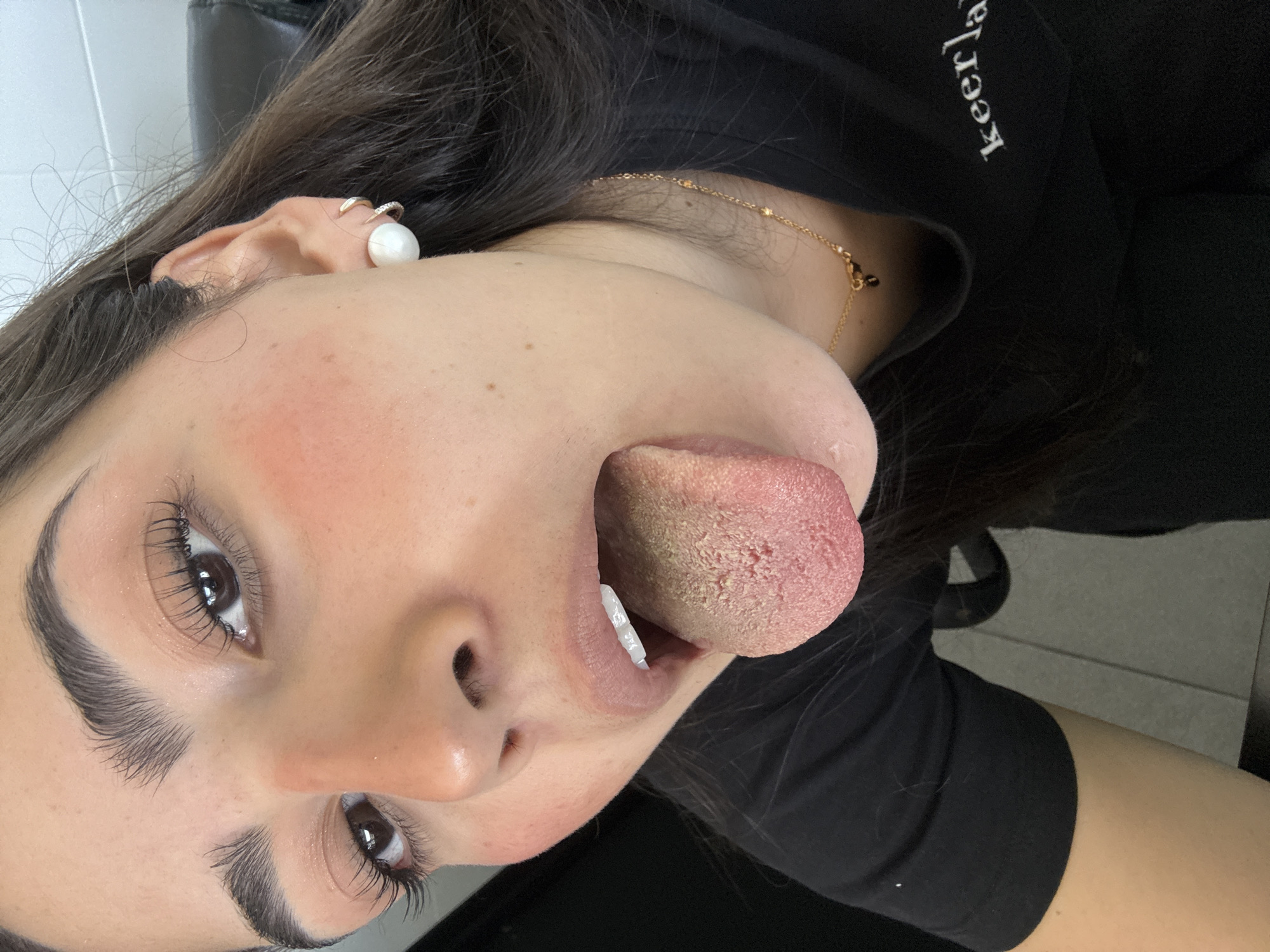 A close-up portrait image, rotated 90 degrees counter-clockwise, depicts a person, likely female, with dark hair and a playful expression, extending their tongue.
The subject's face is prominent, featuring well-groomed, dark eyebrows and dark eyes with long, dark eyelashes. Their left eye is clearly visible, reflecting light. The skin tone is light, with a hint of pink blush on the cheeks. The lips are slightly parted, revealing white teeth on the lower jaw. The tongue is fully extended from the mouth, appearing wide and thick. Its surface displays a prominent, rough, and somewhat textured coating, predominantly a yellowish-white color towards the back, gradually becoming pinker towards the tip.
Long, dark brown hair frames the subject's face and extends behind her. In her left ear, a large, round, white pearl earring is visible, along with a smaller, gold-colored hoop earring higher on the helix. A delicate gold-colored chain necklace with small beads is worn around the neck. The subject is wearing a black t-shirt, on which the white text "keerla" is partially visible on the upper right side (as oriented in the original image).
The background is indistinct, showing a mix of dark and light areas, suggesting an indoor setting with artificial lighting, possibly featuring a white wall and dark furniture. The location context provided (San Bernardino Tlaxcalancingo, Mexico) is not discernibly evident from the visual cues within the image.
