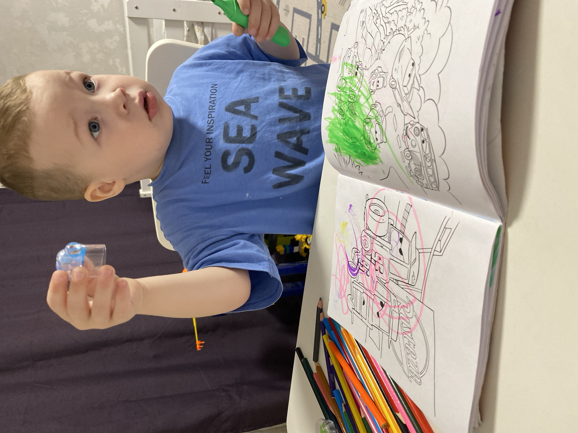 A young boy with fair skin and blonde hair is seated at a white table, engrossed in coloring. He is wearing a blue t-shirt with the words "SEA WAVE FEEL YOUR INSPIRATION" printed on it. In his left hand, he holds a small, clear plastic object, and in his right hand, he holds a green marker.
In front of the boy is an open coloring book. The pages are filled with illustrations, some of which have been colored with green, pink, and other colors. A collection of colored pencils is scattered near the bottom right of the book.
The background features a dark curtain or drape, a white chair, and a wall. The scene appears to be indoors, possibly in a home or daycare setting. There is no information to suggest this image was taken in Sochi, Russia.