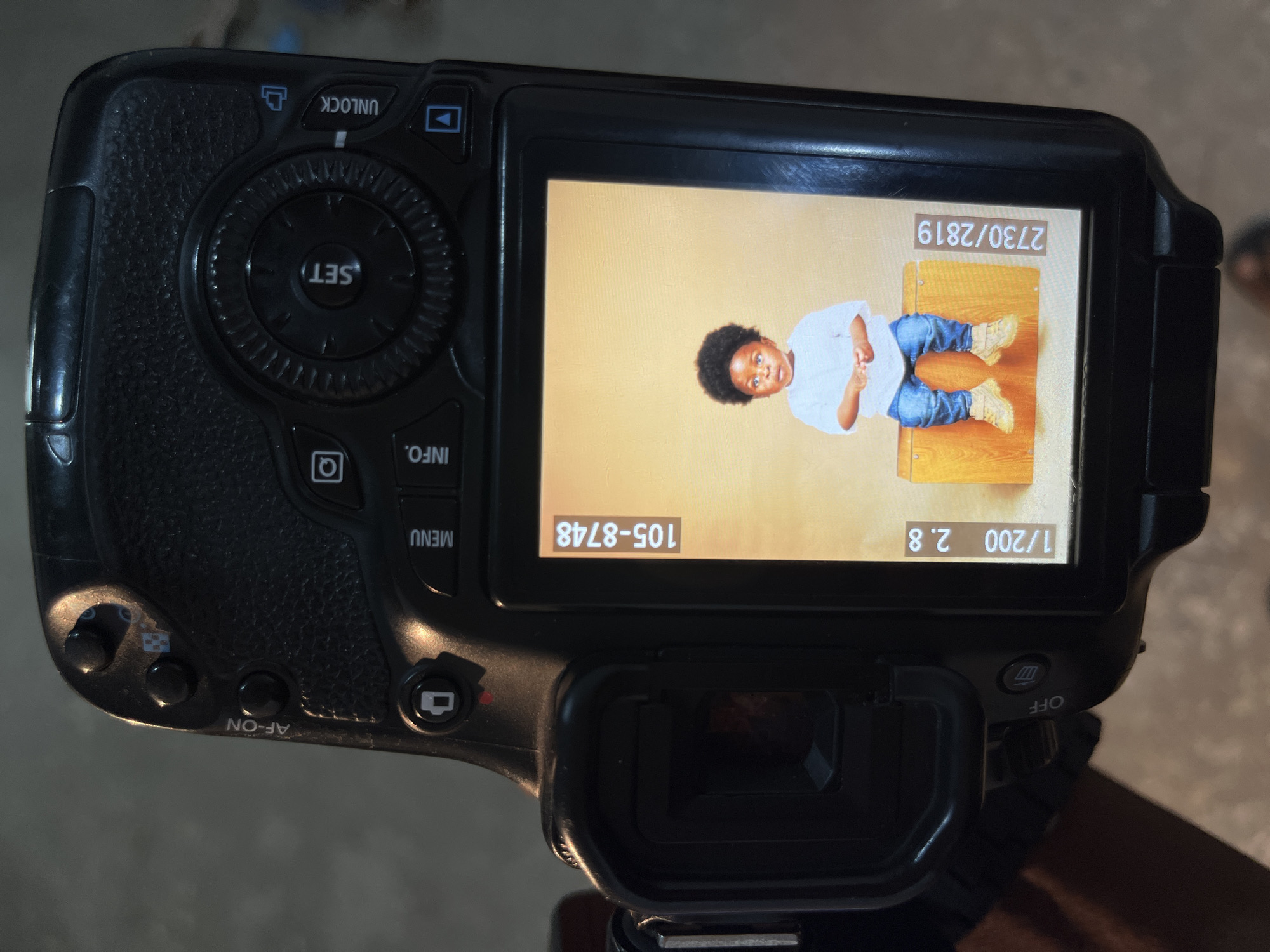 This media file shows a close-up, overhead view of a digital camera displaying a photograph of a young child. The child, with dark curly hair and brown skin, is wearing a white long-sleeved shirt and blue jeans. They are sitting on a wooden surface, looking towards the camera with a slight smile. The background behind the child is a plain, light orange color.
On the camera's LCD screen, there are several pieces of visible text, including "2730/2819", "105-8748", and "1/200 2.8". These appear to be image metadata such as file number, exposure settings, and possibly a date.
The camera itself is black and has various buttons and dials. Visible controls include "UNLOCK", "SET", "INFO.", and "MENU". The "AF-ON" button is also clearly visible, as is the power switch labeled "OFF". The overall scene appears to be indoors, given the lighting conditions and the presence of the camera equipment. The location context provided is Gura Top, Nigeria. The photograph displayed on the screen captures a moment of a child posing for a picture, suggesting a photography session is in progress. The emotions conveyed by the child's expression are neutral to happy.