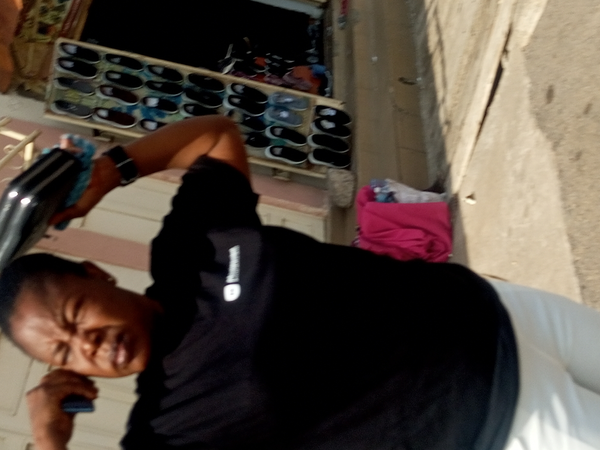 A low-angle shot captures a person in the foreground wearing a black t-shirt and white pants, holding a blue object in their left hand while their right hand is extended and touching their ear. The person's face is contorted, and their eyes are closed, suggesting discomfort or strain. In the background, a shelf displays an assortment of shoes. To the right of the person, a bright pink garment is visible. The scene appears to be outdoors, with a concrete wall on the right and the suggestion of a building on the left, bathed in natural light.