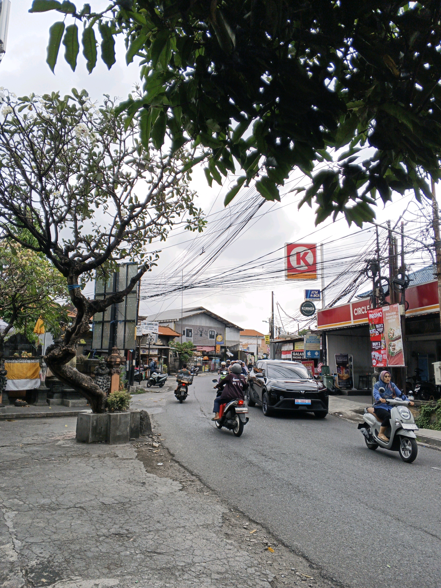 The image captures a street scene in Denpasar, Indonesia, on a cloudy day. In the foreground, a street with cracked asphalt leads into the distance. A mature, gnarled tree with visible branches and some white flowers stands on the left side of the road, rooted in a concrete planter. The tree is adorned with a blue ribbon tied around one of its branches. Behind the tree, there are traditional Balinese stone carvings and a yellow cloth draped over a shrine-like structure.
The road is populated with various vehicles, primarily motorcycles. Several individuals are riding motorcycles, with some wearing helmets and casual attire. A black SUV is also present on the road, with a license plate visible. On the right side of the road, there are shops and commercial establishments. A red and white Circle K convenience store is prominent, with advertising posters displayed outside. Another red sign with a white "K" in a square is visible higher up, also indicative of the Circle K brand. Signs for ATM and BCA are also present. A woman wearing a blue hijab and sunglasses rides a light gray scooter on the right side of the road, while other motorcycles and a few pedestrians are dispersed throughout the scene. Overhead, a dense network of electrical wires crisscrosses the sky, characteristic of urban Indonesian infrastructure. The background reveals more buildings and the overcast sky.
