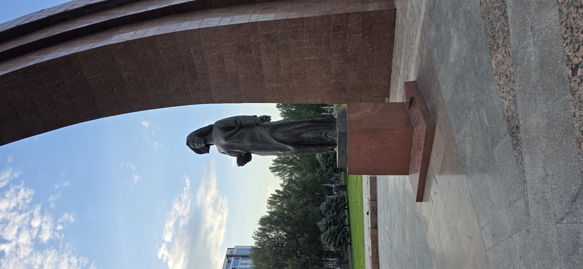 A bronze statue of a woman standing on a pink granite pedestal is the main subject of the image, located in Bishkek City, Kyrgyzstan. The statue depicts a woman wearing a long, flowing robe and holding a small bowl in her left hand. She has dark hair and is looking towards her left. The pedestal is rectangular with beveled edges and sits on a larger, stepped base. Behind the statue, a large, curved structure with a reddish-brown, granite-like surface dominates the right side of the frame. This structure appears to be part of an archway or monument.
In the background, to the left of the statue, is a modern, multi-story building with glass windows. A line of green trees, including some pine trees, forms a backdrop behind the statue and extends across the middle ground. The ground in the foreground and around the statue is paved with large, square granite tiles in varying shades of gray and pink. The sky above is blue with scattered white clouds. The overall scene suggests an outdoor public space, likely a memorial or civic area.