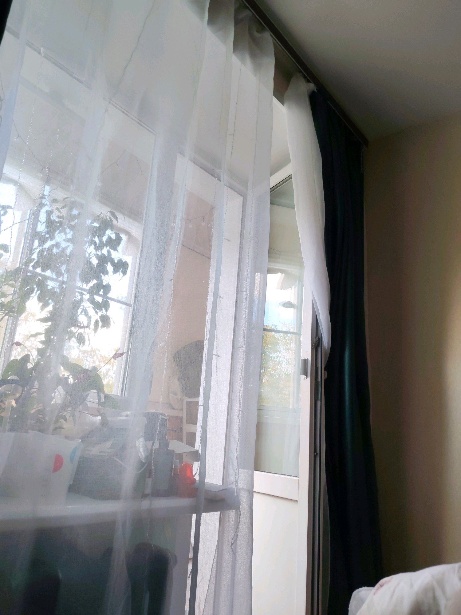 A view through a sheer white curtain in a residential setting, likely a balcony or large window, reveals a plant on a windowsill with several bottles behind it. The curtains are hung from a track on the ceiling and extend down, partially obscuring the view outside. A dark green curtain is visible to the right, drawn partially closed. The overall impression is one of an indoor space with natural light filtering in. The location is specified as Izhevsk, Russia.