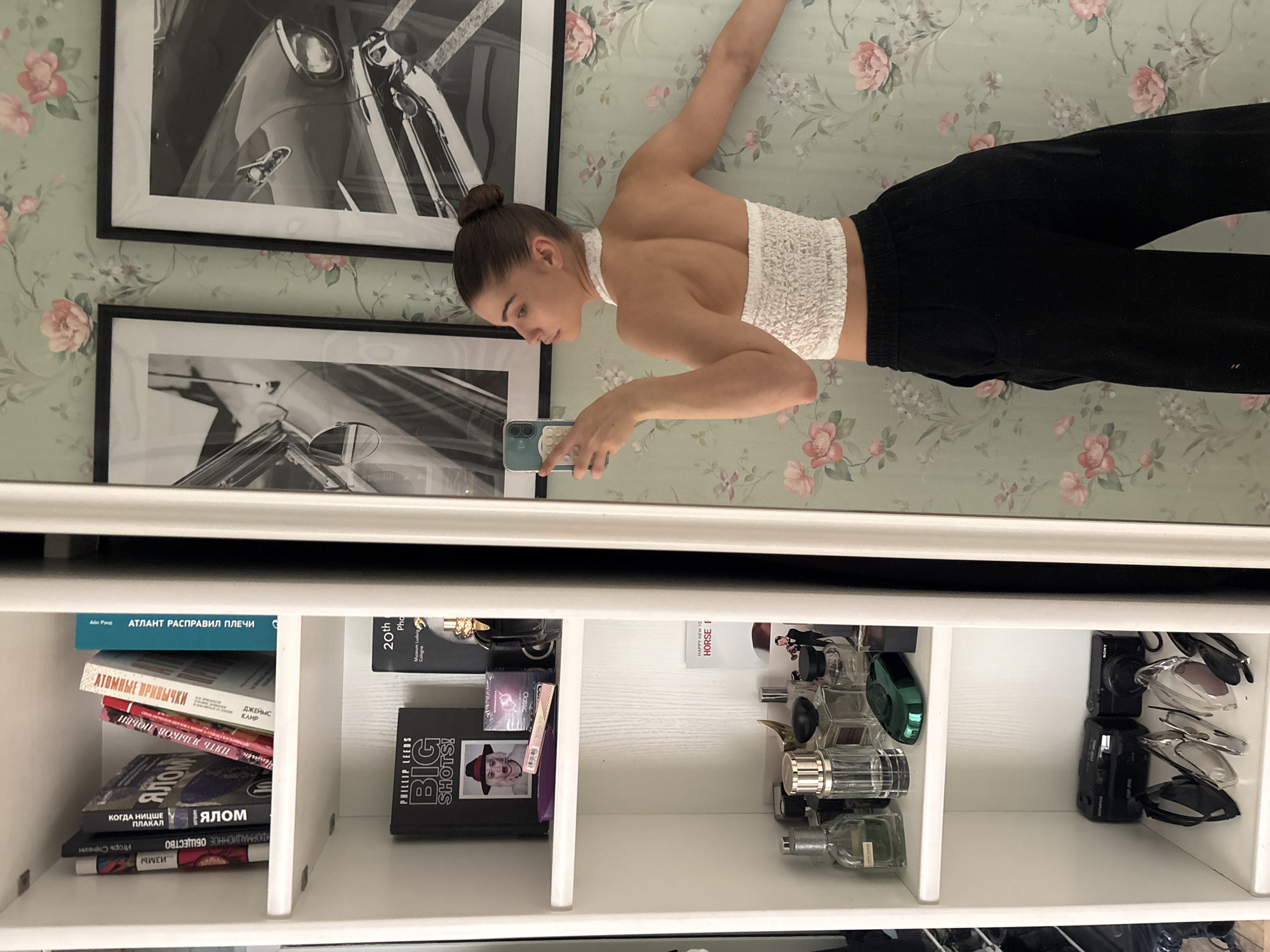 A young woman is reflected in a mirror above a white shelving unit. She has brown hair in a bun and is wearing a white halter top and black pants. She is holding a smartphone up, presumably taking a picture of herself in the mirror. Behind her are two framed black and white photos with floral wallpaper as a backdrop. The shelves contain a variety of items, including books, perfumes, cameras, and sunglasses. The titles of some of the books in Russian are visible. The location appears to be a bedroom or dressing area, but not explicitly Moscow.