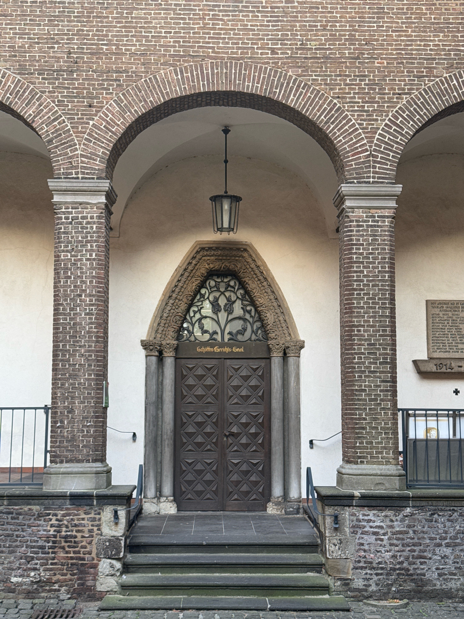 The image shows the entrance to the St. Martin's Church in Cleves, Germany. The entrance features a large, ornate wooden door set within a stone archway. Above the door is a decorative wrought-iron window with a floral pattern. The entrance is flanked by two large brick columns and is approached by a set of stone steps. The building itself is constructed of red brick, with a series of arches visible in the background. There is a hanging lantern above the entrance. To the right of the entrance, a plaque with text is mounted on the wall, along with the year "1914". A cross is visible below the plaque. A metal railing runs along the sides of the entrance area. The ground in front of the steps is paved with cobblestones. The scene is viewed from a low angle, looking up at the entrance.
