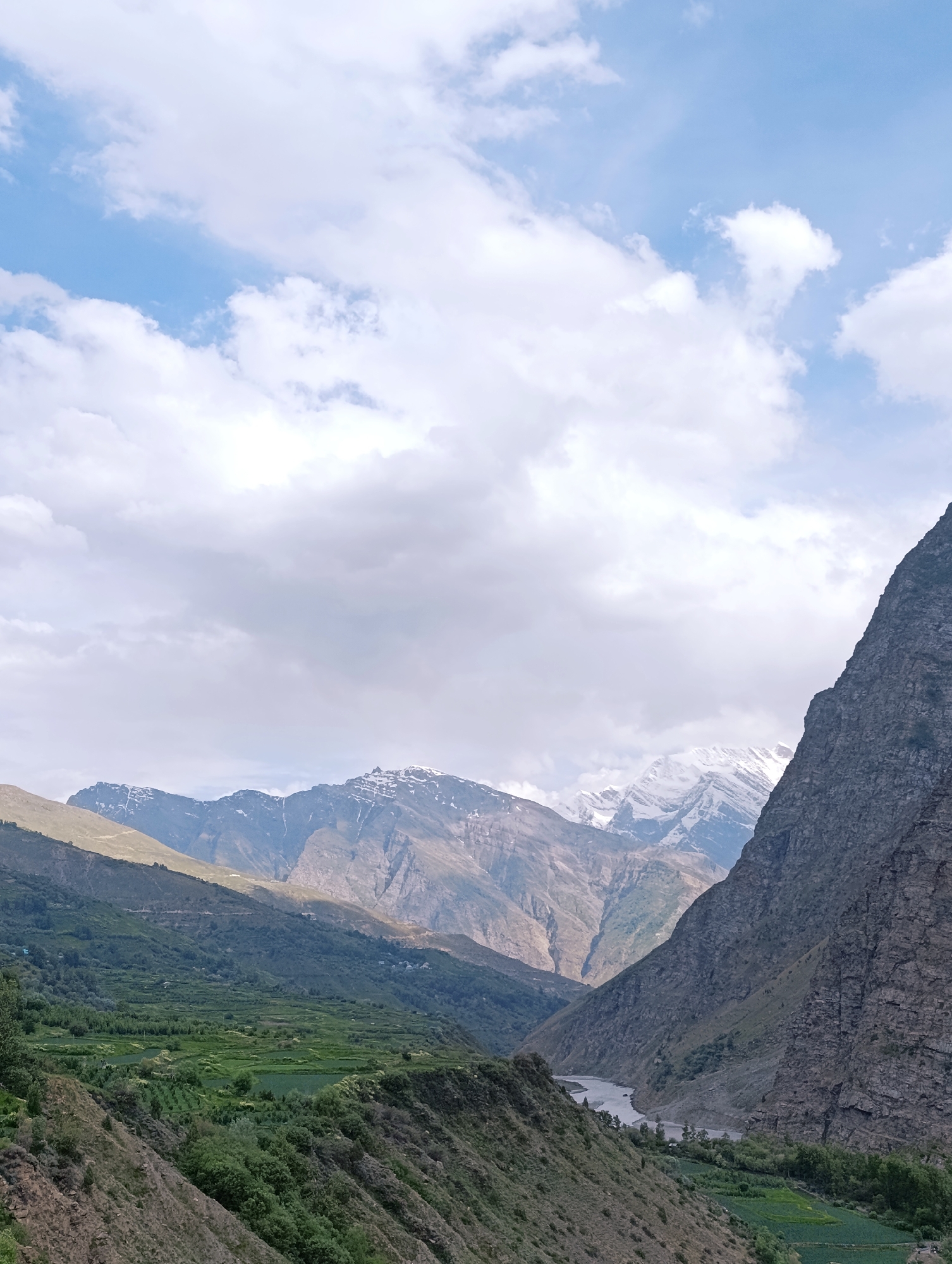 The image depicts a valley in the vicinity of Kirting, India. A river flows through the valley, flanked by steep, rocky mountains on the right and gentler, terraced slopes on the left. The terraced slopes are covered in lush green vegetation, suggesting agricultural activity. In the background, majestic, snow-capped mountains rise against a sky filled with dramatic clouds. The overall scene conveys a sense of natural grandeur and tranquility. There are no people or animals visible in the image.