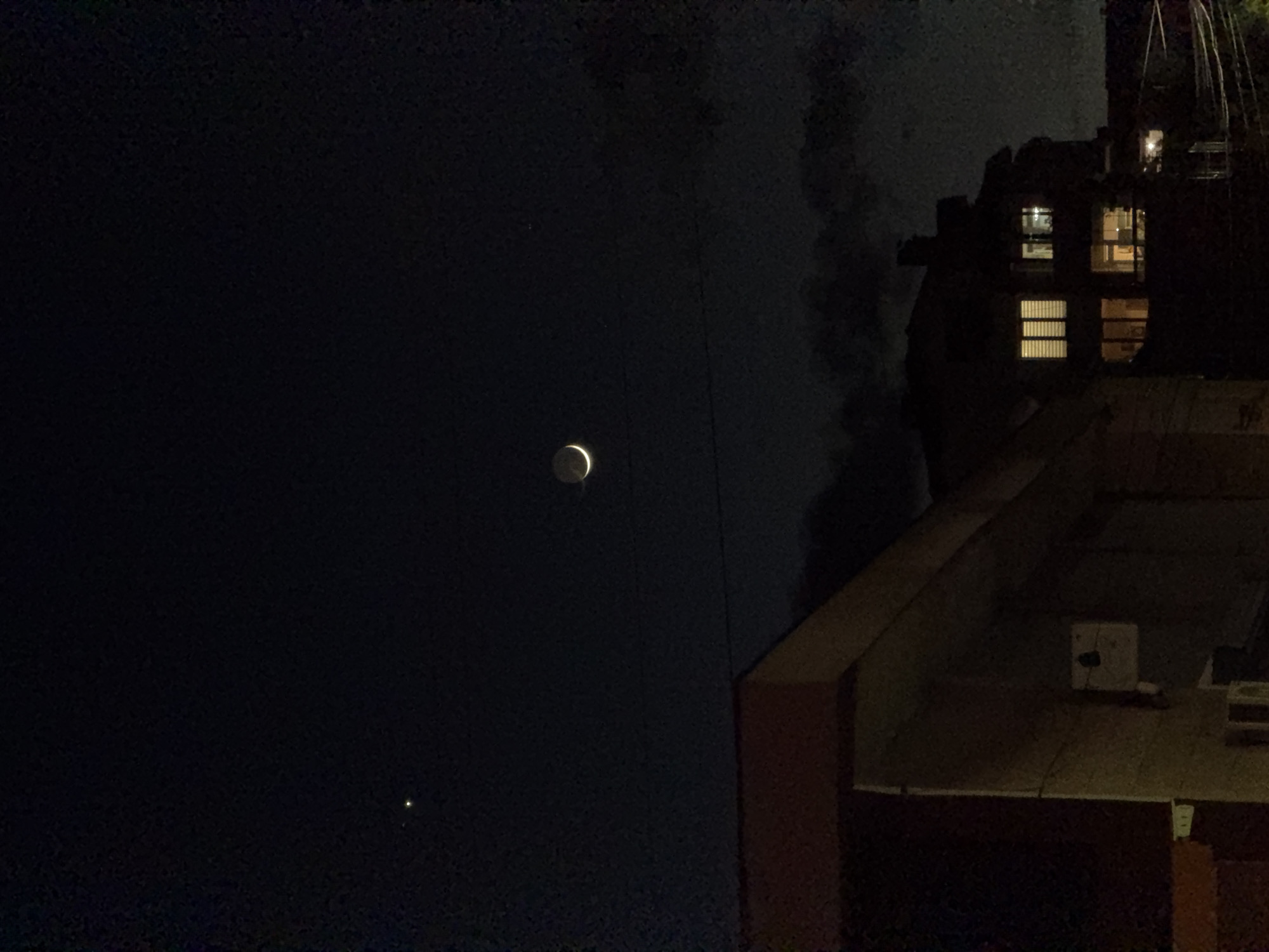 The image shows a night scene with a crescent moon and a bright star or planet visible in the dark sky. A building with lit windows is on the right side of the frame, and the roof of a lower building is visible in the foreground. The scene appears to be in an urban environment. There is nothing in this image that indicates that the image was taken in Burgas, Bulgaria.