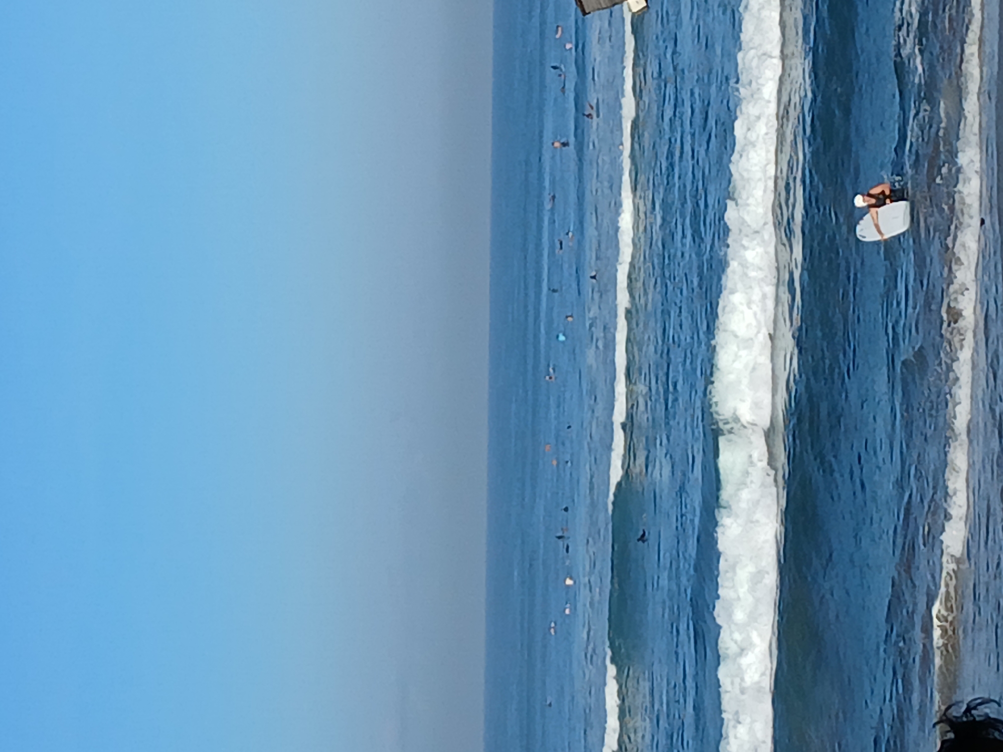 A wide shot captures a serene ocean scene with a clear blue sky. Several surfers are dispersed across the water, with some paddling and others waiting for waves. The foreground shows a person in the water, holding a white surfboard and wearing a white cap and dark top. Gentle waves are rolling towards the shore. The environment is an ocean setting, consistent with the beaches of North Kuta, Indonesia.
