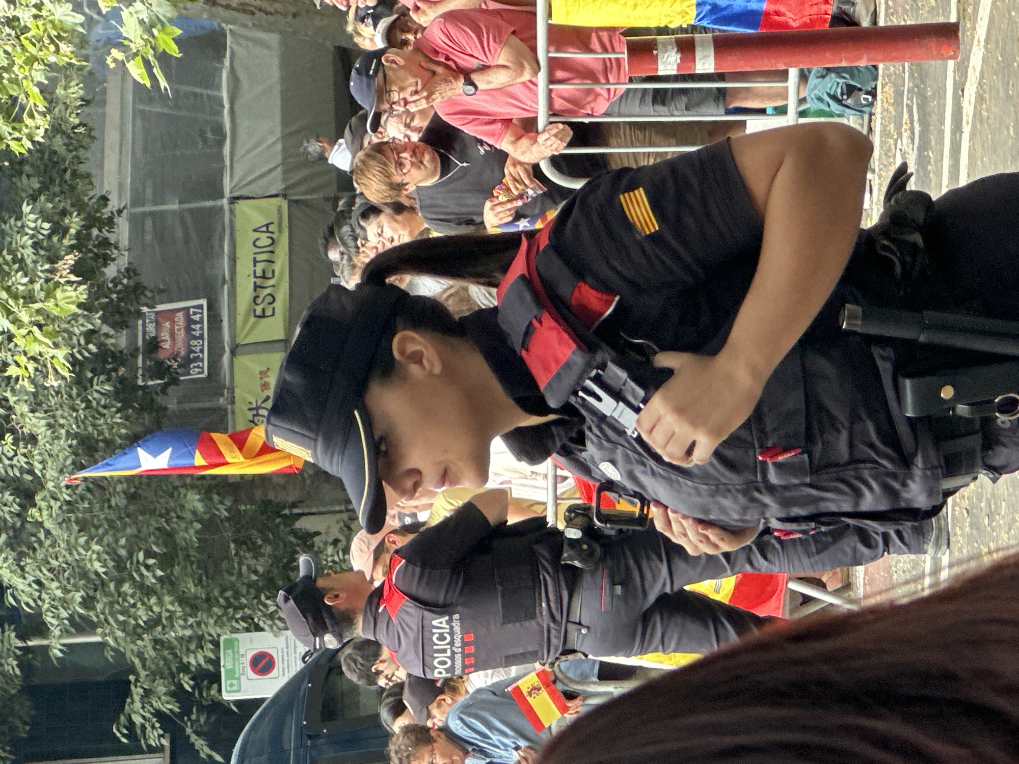 The image captures an outdoor scene in what appears to be a public gathering or event. In the foreground, a female police officer in uniform is prominently featured, facing to her right in profile. She is wearing a black cap, a black tactical vest with red accents, and dark pants. The vest has "POLICIA" printed on the back in white lettering, along with other insignia. A gun holster is attached to her belt. To her left, another police officer in a similar uniform is visible from the waist up, also facing away from the camera. He is holding a Spanish flag.
In the background, a diverse crowd of people are gathered. Some individuals are holding flags, including what appears to be a Catalan flag hanging from a pole and another Spanish flag. Several banners are visible in the background, one with the word "ESTÈTICA" in yellow text on a brown background. Trees and foliage overhang the scene, suggesting an urban park or street setting. The overall atmosphere appears to be one of a public demonstration or parade. The lighting suggests it is daytime.