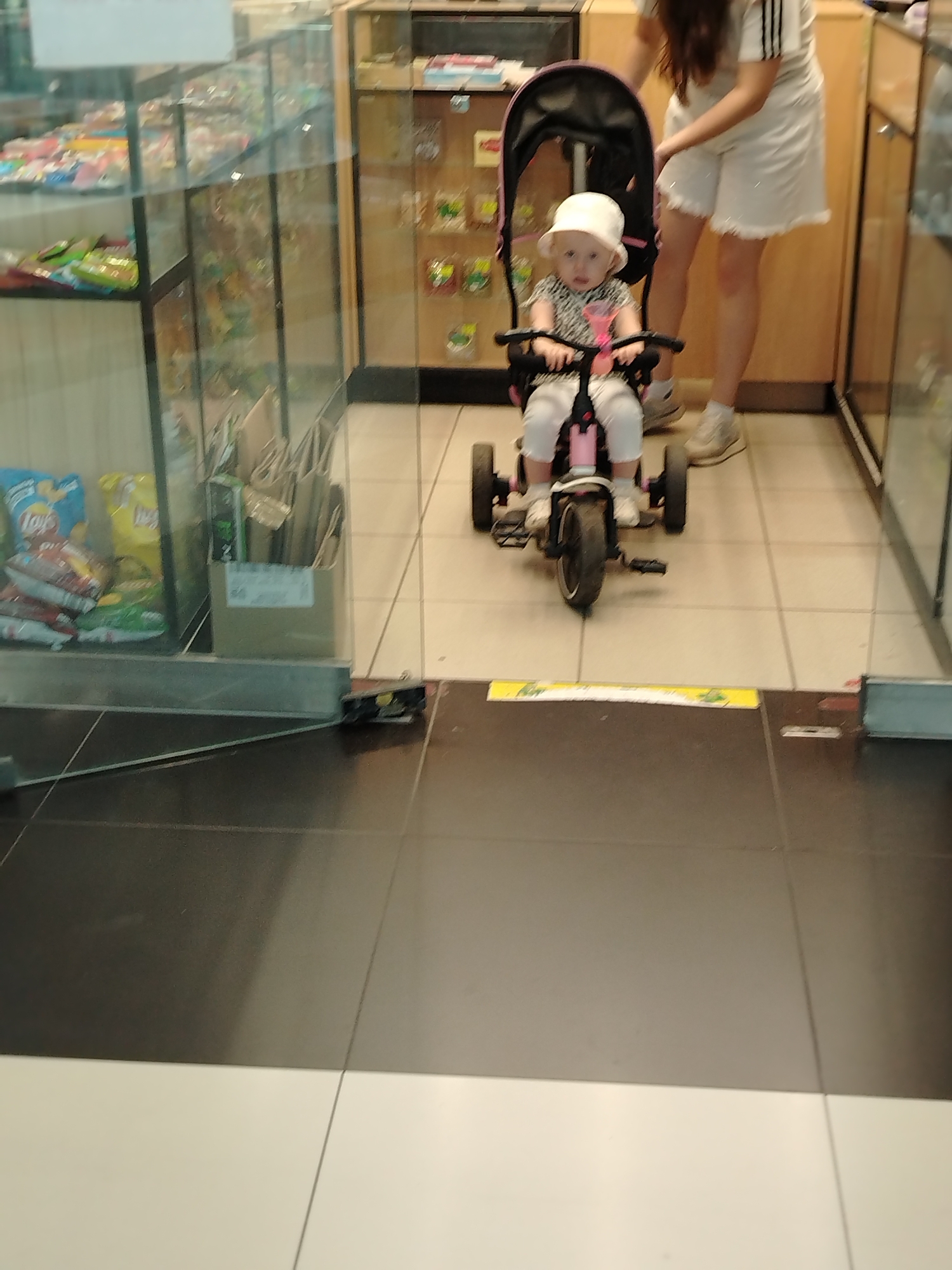 In a brightly lit indoor commercial space, a young child wearing a white bucket hat and a black and white patterned shirt sits in a pink tricycle with a black canopy. The child's legs are extended and appear to be resting on the pedals. To the right of the tricycle, a person with long brown hair, wearing a white t-shirt with black stripes and white shorts, stands partially obscured from view, their hands near the tricycle's canopy. The background features glass display shelves filled with various items, suggesting a retail environment such as a convenience store or small market. The floor is tiled with a pattern of alternating black and white squares. The scene is consistent with a shopping location within the city of Izhevsk, Russia.