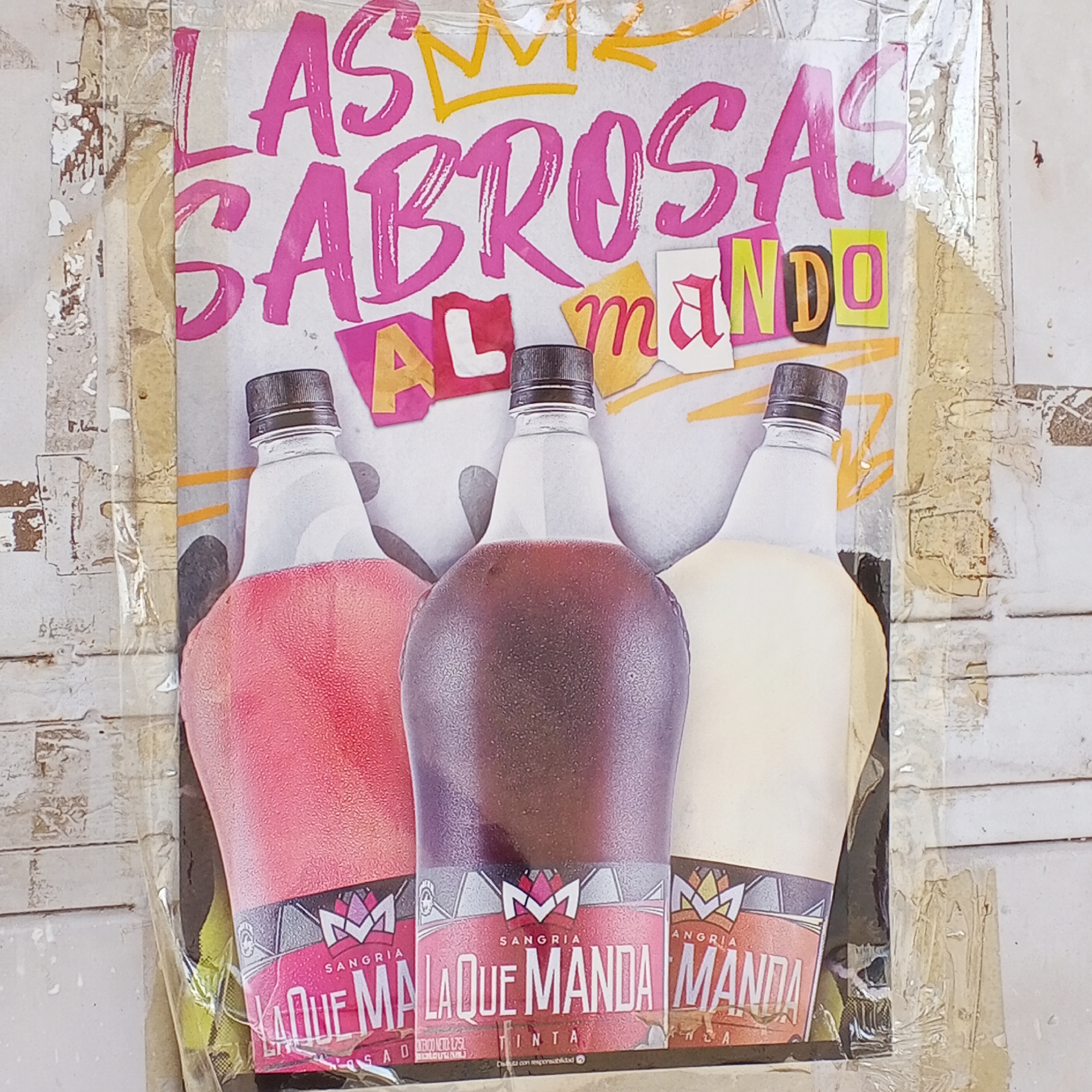 A colorful advertisement poster, possibly for a beverage brand, is shown affixed to a textured, weathered surface, likely a wall. The poster features three bottles arranged side-by-side. The liquid inside the left bottle is a reddish-pink, the middle bottle contains a dark reddish-brown liquid, and the right bottle holds a creamy, pale yellow liquid. All bottles have black caps and appear frosty, suggesting they are cold drinks.
Above the bottles, large, vibrant pink letters spell out "LAS SABROSAS". Below this, in a mixed collage-style font of pink and yellow cut-out letters, is "AL mando", with a yellow crown-like drawing above the text. This forms the slogan "LAS SABROSAS AL mando," which translates to "The tasty ones in charge" or "The flavorful ones taking command."
Each bottle has a label at its base, featuring a stylized "M" logo. The visible text on the labels confirms the product is "SANGRIA" and prominently displays "LA QUE MANDA" (The one that commands). The middle bottle's label explicitly states "SANGRIA LA QUE MANDA TINTA" (red/dark sangria), while the left one shows "LA QUE MA" (likely "LA QUE MANDA ROSADA" for pink/rosé sangria). The right bottle's label is partially obscured but shows "MANDA". Smaller text on the labels includes "CONT. NETO 1.75L" (net content 1.75L) and "Disfruta con responsabilidad" (Enjoy responsibly).
The background surface to which the poster is attached is light-colored and shows signs of wear, with peeling paint or material visible along the edges of the poster, particularly on the left and right sides. Clear tape or plastic sheeting appears to be used to secure the poster, reflecting light in places. The overall impression is that of an informal, perhaps street-side, advertisement.