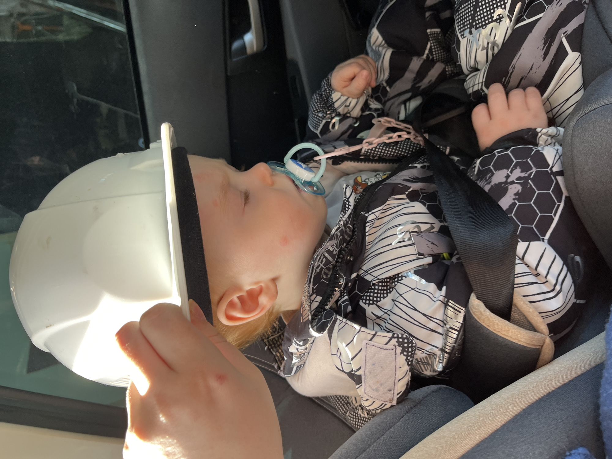 The image displays an interior view of a vehicle in the city of Chaikovsky, Russia. A light-skinned hand is positioning a white hard hat on the head of a young child. The child, a toddler with light-colored hair, is seated in a dark-colored car seat and appears to be resting with eyes closed. A light blue and white pacifier is in the child's mouth, connected by a pink plastic chain. Red marks are visible on the child's left cheek and above their left ear. The child is wearing a black, white, and grey patterned winter jacket, secured by a dark safety harness with padded straps. The vehicle's dark grey interior is visible in the background, including a silver door handle to the right and a window reflecting a faint green object. A beige seatbelt and a portion of a blue fabric object are visible on the left side of the frame.