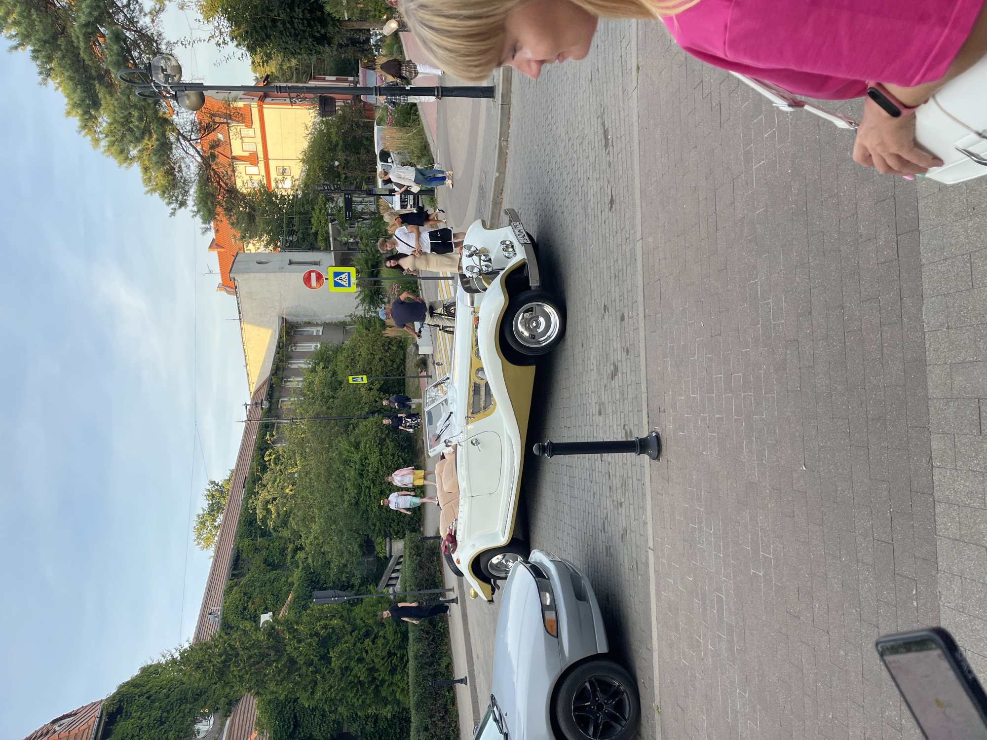 On a cobblestone street in Svetlogorsk, Russia, a vintage white and gold convertible automobile is parked. A silver sedan is parked to its left. Several people are walking on the sidewalk and across the street. Buildings line the street, some covered in ivy. Streetlights and trees are also visible. The sky is clear and blue.