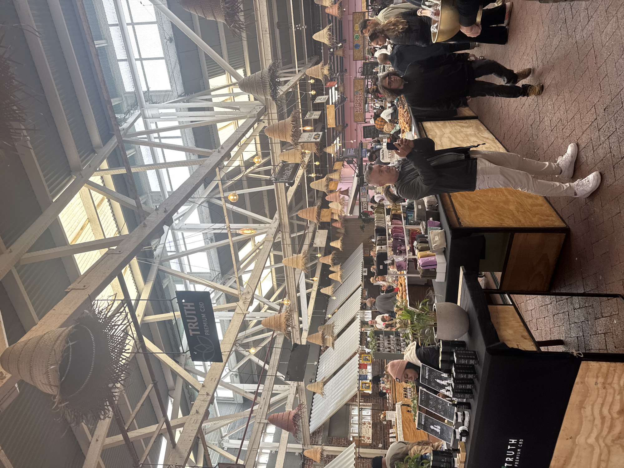 The image shows the interior of the Old Biscuit Mill market in Cape Town, South Africa. The high ceiling is exposed, revealing a complex network of wooden beams and skylights. Numerous woven baskets and light fixtures hang from the structure.
Market stalls are set up on either side of a brick pathway. A man in white pants and sneakers, wearing a jacket, is photographing a stall with a mobile phone. Another person with long hair walks nearby. Several other people are browsing the stalls.
One stall has a black tablecloth with "TRUTH PREMIUM CBD" printed on it. The stall displays items like creams and lotions, along with informational signs reading "BENEFITS." Other visible stalls have goods such as hats and bags.