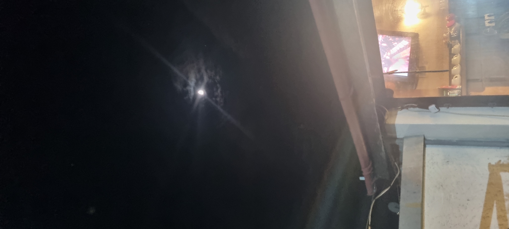 A nighttime exterior view shows the moon in a dark sky visible above a building. The building has a weathered appearance, with visible pipes, wiring, and a partially obscured painted word. Inside the building, a television screen is visible, surrounded by various objects on shelves. The specific location in the city of Unknown, Unknown cannot be determined from this image.