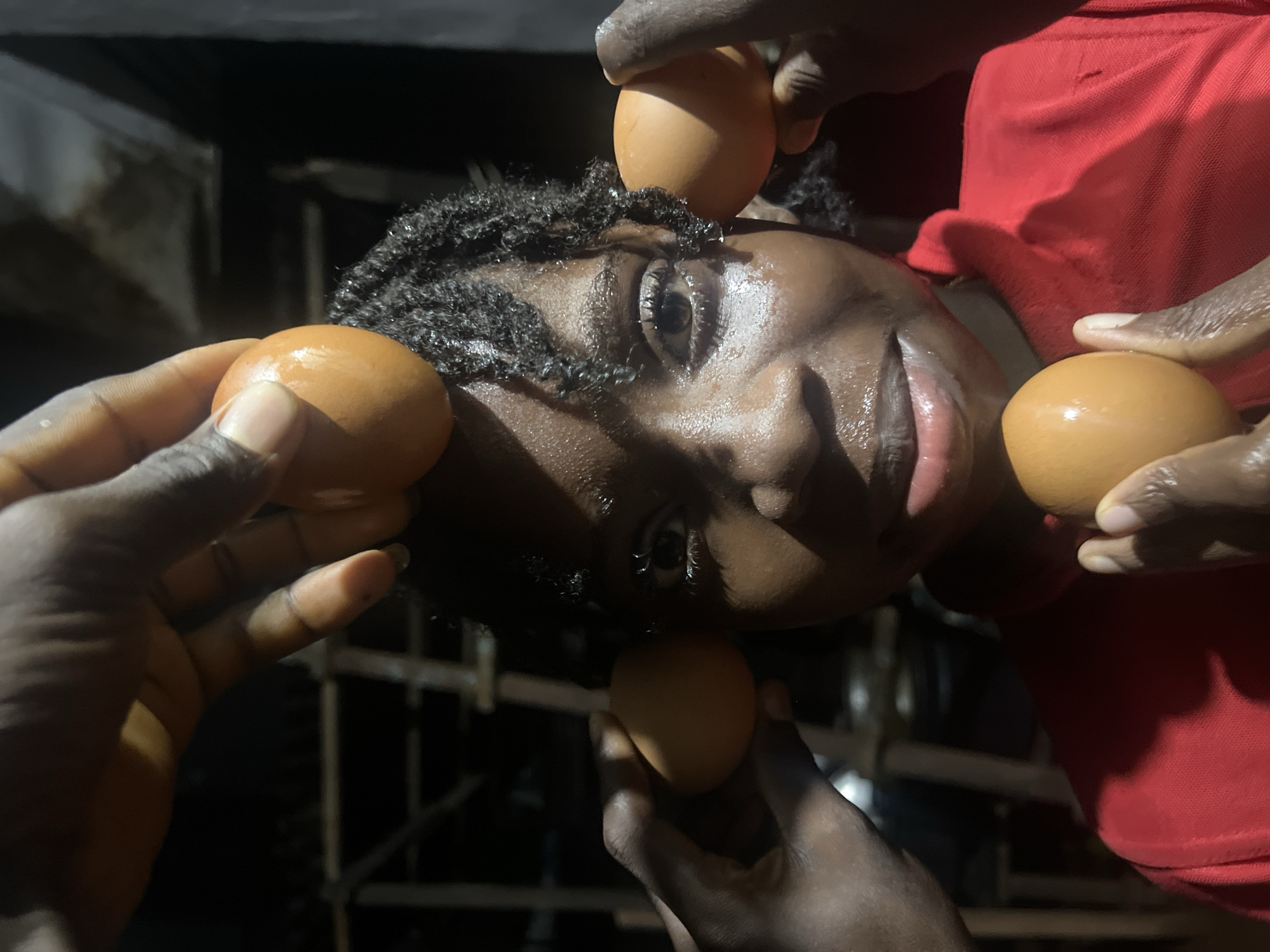In Nkpor, Nigeria, a close-up shot captures a young Black person with braided hair, their face tilted upwards, and a bright red shirt. Their skin glistens with sweat. Multiple brown eggs are positioned around their head and chin, held by several hands. One egg rests atop their head, two are placed on either side of their temples, and another is held in front of their chin. The background is dark and out of focus, with hints of scaffolding or a building structure.