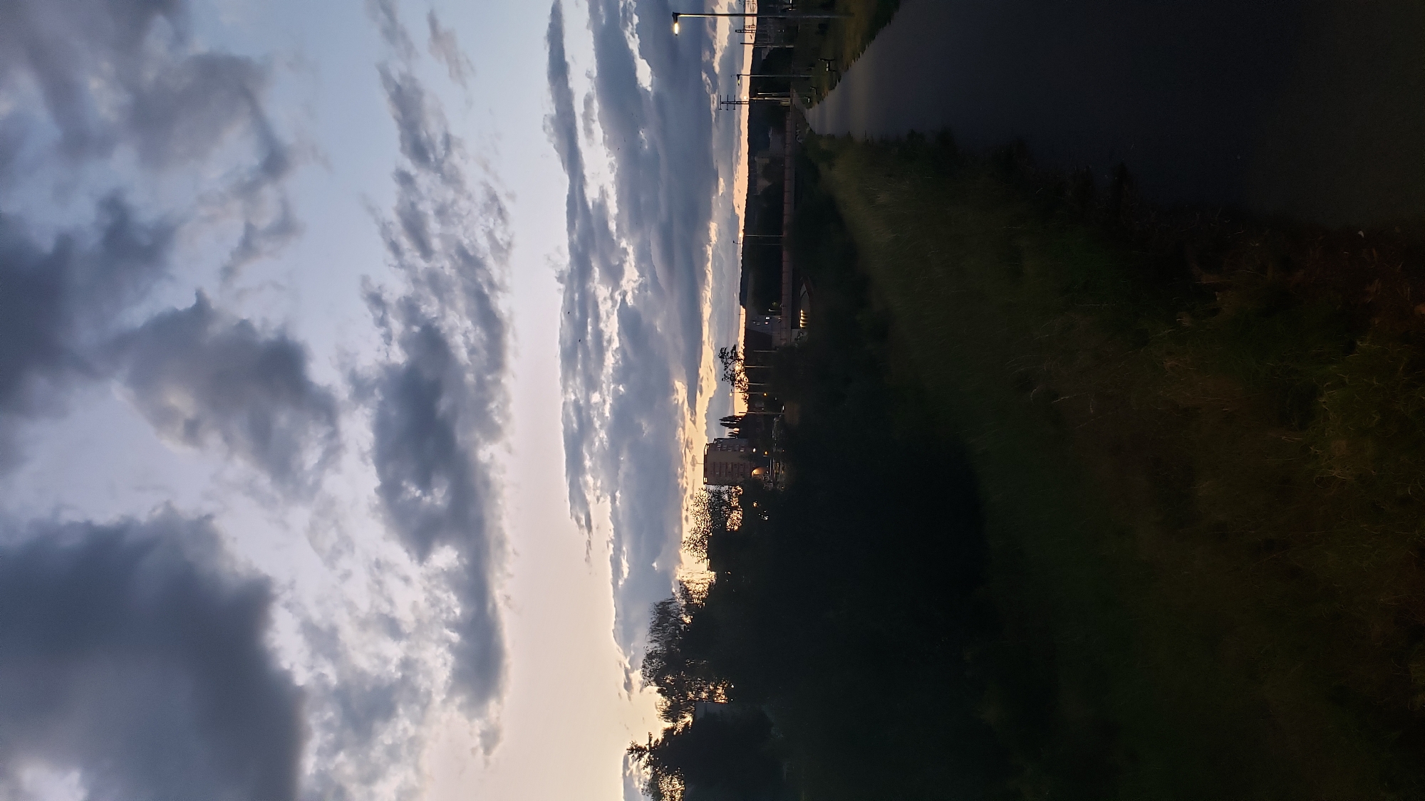 The image captures a dusky scene along a paved pathway in Prešov, Slovakia, likely near the Torysa River. The sky is filled with dramatic, dark clouds interspersed with lighter, purplish hues near the horizon, suggesting either sunrise or sunset. Silhouetted against the sky are trees and buildings, including a prominent multi-story structure. The pathway itself is dark and appears to be an urban or park walkway, with streetlights visible at intervals and benches along its edge. To the left of the pathway, tall grass and foliage are visible, fading into darkness. The overall atmosphere is serene and slightly somber, indicative of the transition between day and night.