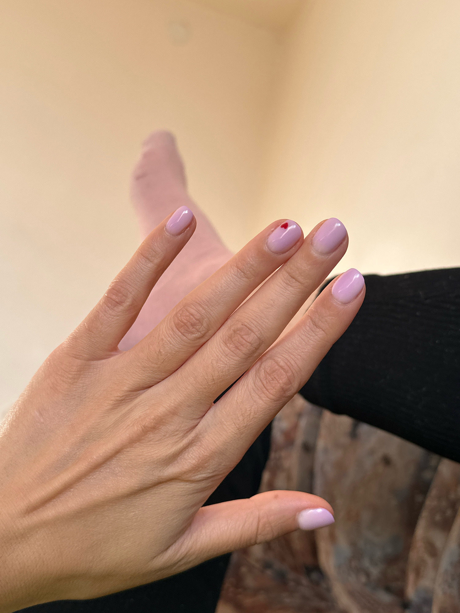 The image shows a close-up of a person's left hand with light purple nail polish. A tiny red heart-shaped design is visible on the thumbnail. The background is a soft, neutral beige. Part of a pink sock or leg is visible in the upper left corner, and a dark, textured fabric, possibly clothing, is in the lower right. The lighting is soft and diffused, suggesting an indoor setting. The image does not provide any information that would indicate a location in Gyumri, Armenia.