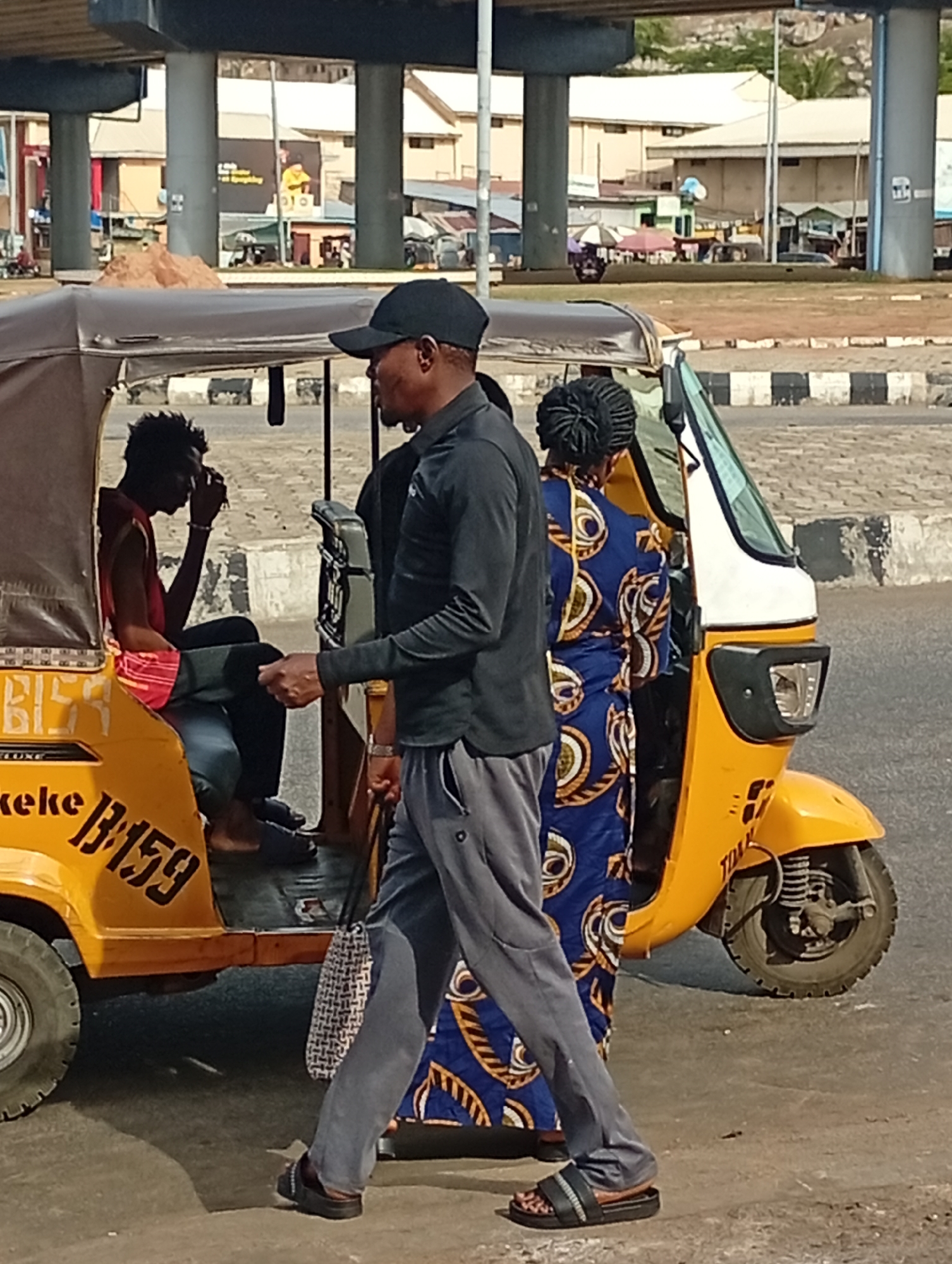The image shows a yellow tricycle, locally known as a "keke," with the number "B-159" printed on its side, at a location in Lokoja, Nigeria, as suggested by the writing on the keke. A man wearing a black cap, long-sleeved black shirt, and grey trousers walks past the keke. A woman wearing a blue and gold patterned dress stands near the entrance. Inside the keke sits a man wearing a red shirt. The tricycle appears to be on a paved road near an urban environment with buildings and an elevated structure in the background.