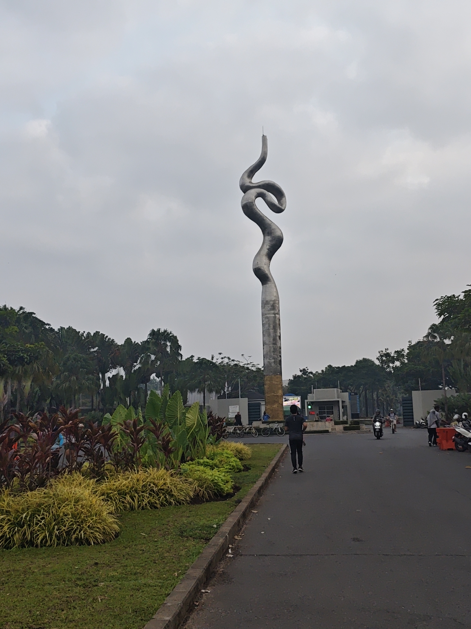 The image depicts a view of the Monument of the Great Serpent (Monumen Ular Tangga) in Bacin, Indonesia, on an overcast day. A tall, abstract, metallic sculpture resembling a coiled serpent dominates the center of the frame. The sculpture is positioned on a paved road leading towards an entrance with a gate and buildings. To the left of the road, there is a well-maintained garden with lush green grass, yellow ornamental plants, and large-leafed plants with reddish-brown foliage.
Several people are visible in the scene. A man in a dark t-shirt and pants is walking away from the camera on the paved road, towards the entrance. In the background, on the road, a person is riding a motorcycle, and further ahead, there are other individuals and motorcycles near the entrance gate. On the far left, near the garden, a few bicycles are parked, and a person is standing beside them.
The environment appears to be an urban park or a landscaped public area, with mature trees forming a backdrop behind the entrance structures. The sky is uniformly grey and cloudy, indicating diffused lighting conditions. The road surface is dark asphalt, and a concrete curb separates the road from the garden area. The overall impression is one of a public space with a prominent artistic landmark.