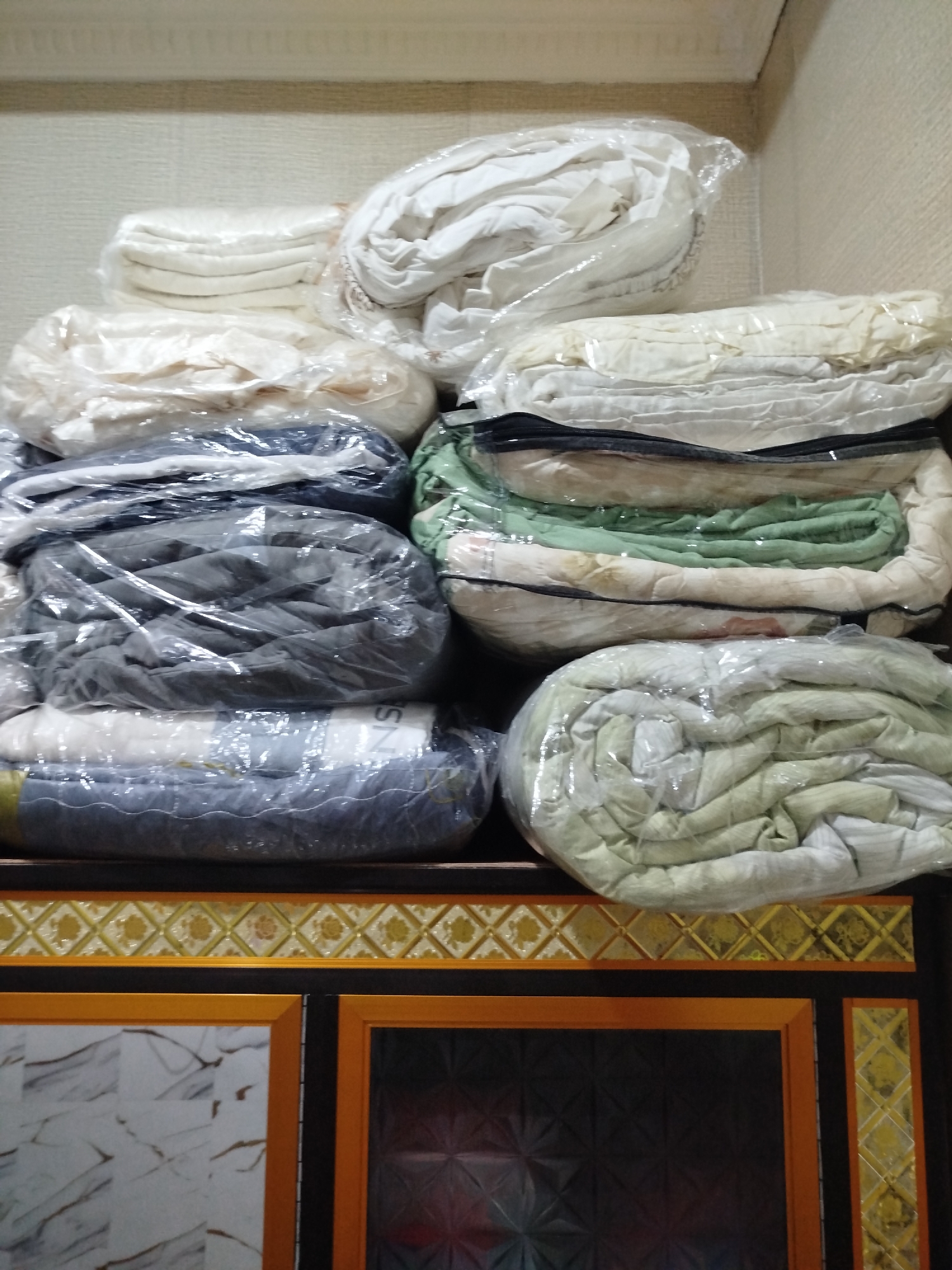 A pile of neatly folded and plastic-wrapped bedding is stored on a shelf above a dark wooden cabinet with decorative trim. The bedding appears to be in various colors, including white, cream, light green, and gray. The background wall is textured and light-colored. The cabinet below has ornate yellow and brown decorative panels and what appear to be dark cabinet doors with a patterned texture. The lighting suggests an indoor setting. There are no people or animals visible in the image. The overall scene is one of organized storage. The location in the city of Bandungrejo, Indonesia is not determinable from the visual information presented.