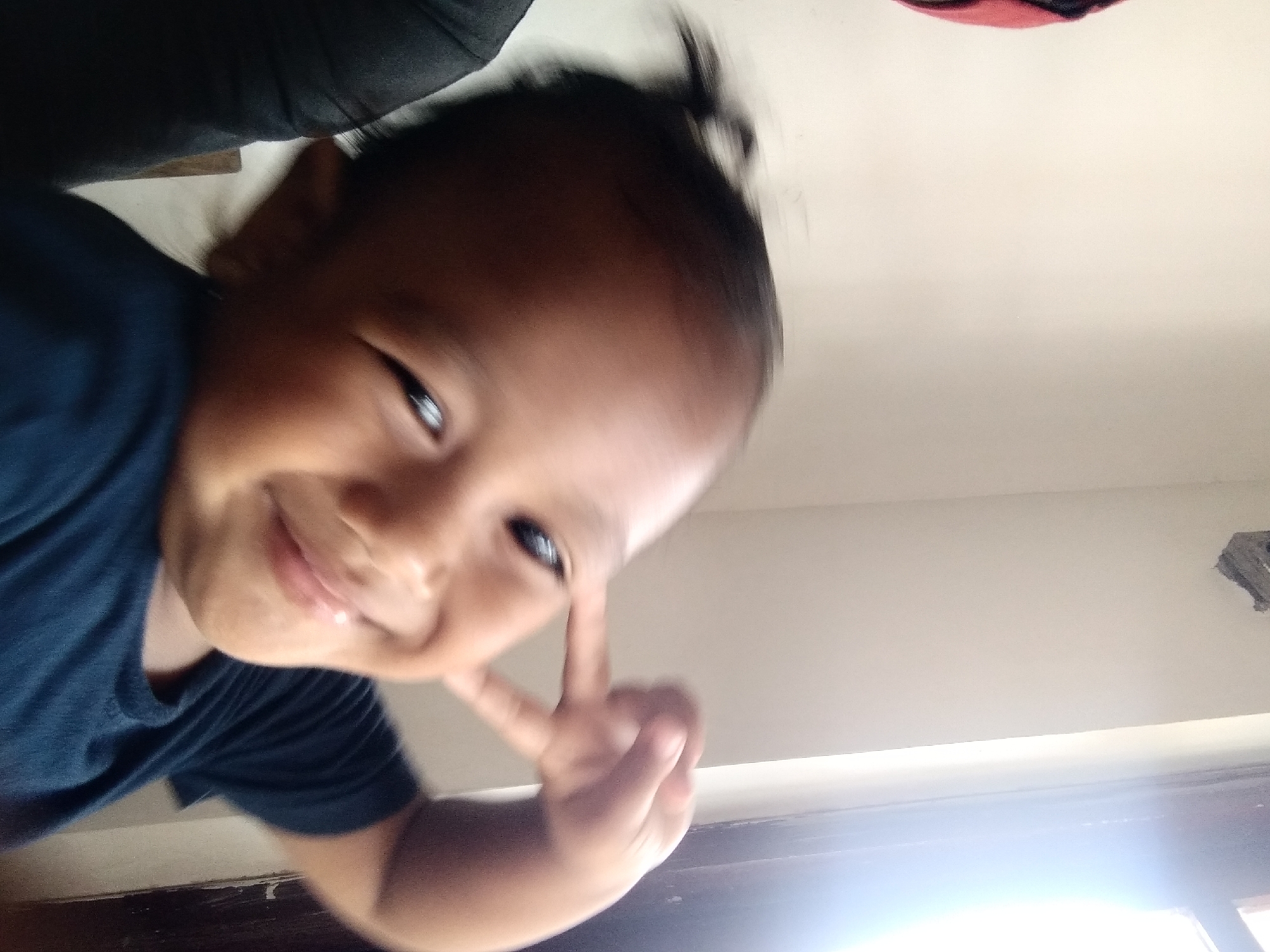 A young child with dark hair pulled into a small ponytail is captured in a close-up, slightly blurry selfie. The child, wearing a dark blue t-shirt, is looking at the camera with a wide smile and has their right hand raised, fingers forming a peace sign near their eye. The background consists of a plain, light-colored wall and a partially visible window with bright light streaming through. The scene appears to be indoors, possibly in a residential setting in Pandak, Indonesia.