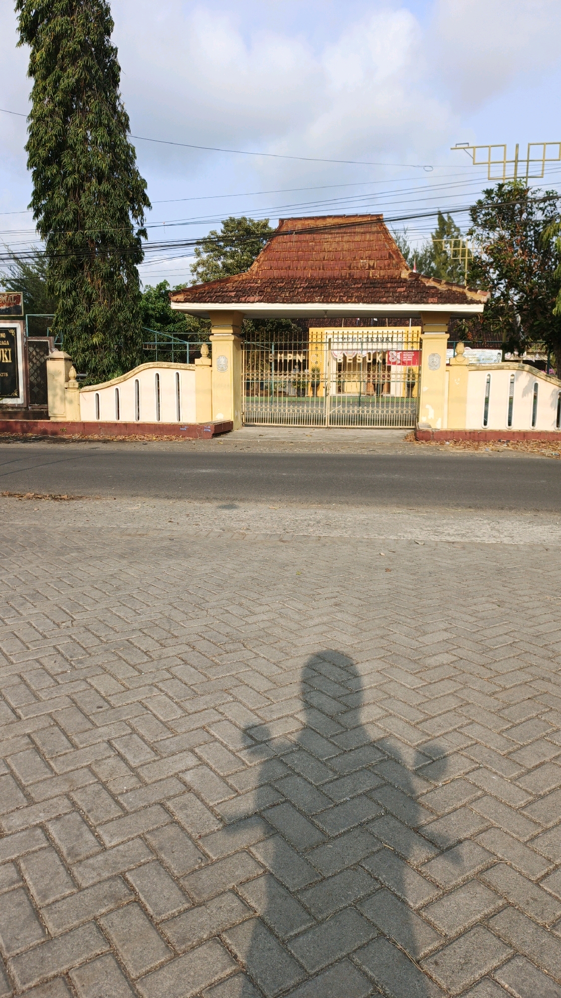 In Tanggulwelahan, Indonesia, a sunlit scene depicts the entrance to a building. The foreground features a brick-patterned walkway with a prominent shadow of a person in the lower right corner, arms outstretched. Beyond the walkway is a paved road. The middle ground showcases a yellow gate adorned with metal bars and a red-tiled roof structure above it. Flanking the gate are beige walls with vertical slits. To the left of the gate, a tall, slender tree with dense green foliage stands prominently. In the background, hints of buildings, trees, and a cloudy sky are visible.