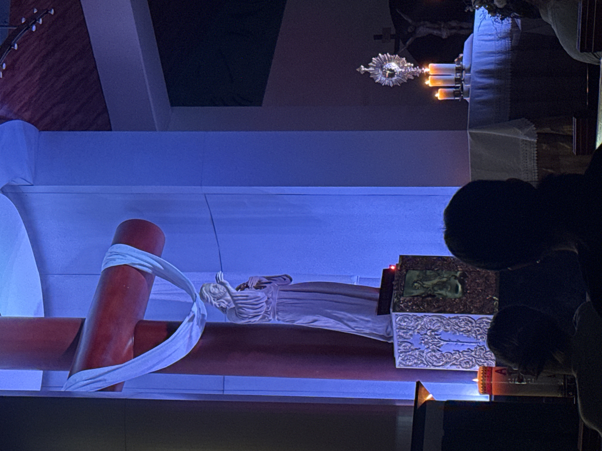 A low-angle, indoor, nighttime shot captures a religious scene at Unknown, Unknown. The composition features a red crucifix with a draped white cloth. A white statue is nearby.
Visible in the background is a monstrance with candles. People are present. The scene is illuminated by both natural and artificial light sources.