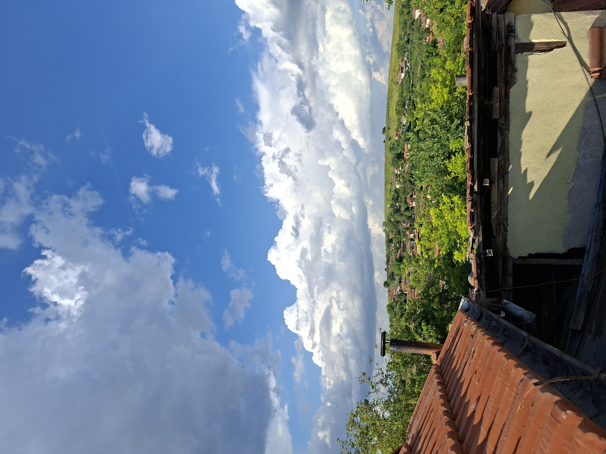 The image shows a view from a rooftop in Bojuritsa, Bulgaria. The foreground consists of a terracotta tile roof with a rusty chimney and a partial view of a dilapidated building structure. Beyond the building, a green landscape dotted with houses extends towards the horizon. The sky occupies a significant portion of the image, featuring a bright blue area with scattered white clouds and a large bank of cumulonimbus clouds dominating the center.