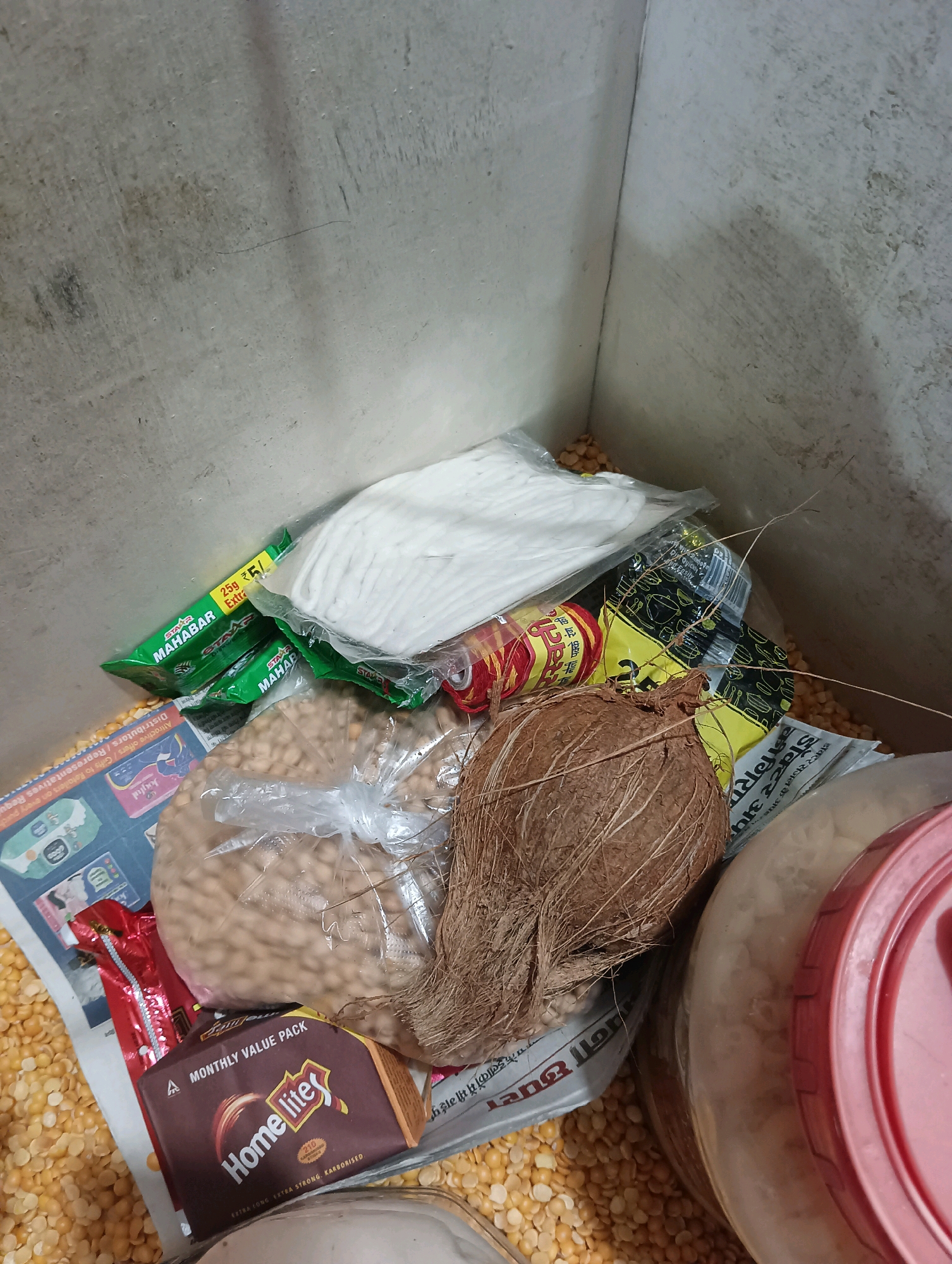 In the city of Dakhin Parsa, India, a concrete storage bin contains various dry goods, including a whole coconut, a bag of chickpeas, and several packages of what appear to be food items. Two green packages labeled "MAHABAR" are visible, alongside a white bundle of material, possibly wicks or cotton. Red and yellow spools of thread are also present. Scattered yellow lentils are seen throughout the bin, along with some loose chickpeas. A partially visible pink-lidded container sits to the right of the image. The bin's interior walls are concrete, showing signs of wear and some staining. The scene depicts a collection of everyday items stored within a utilitarian container.