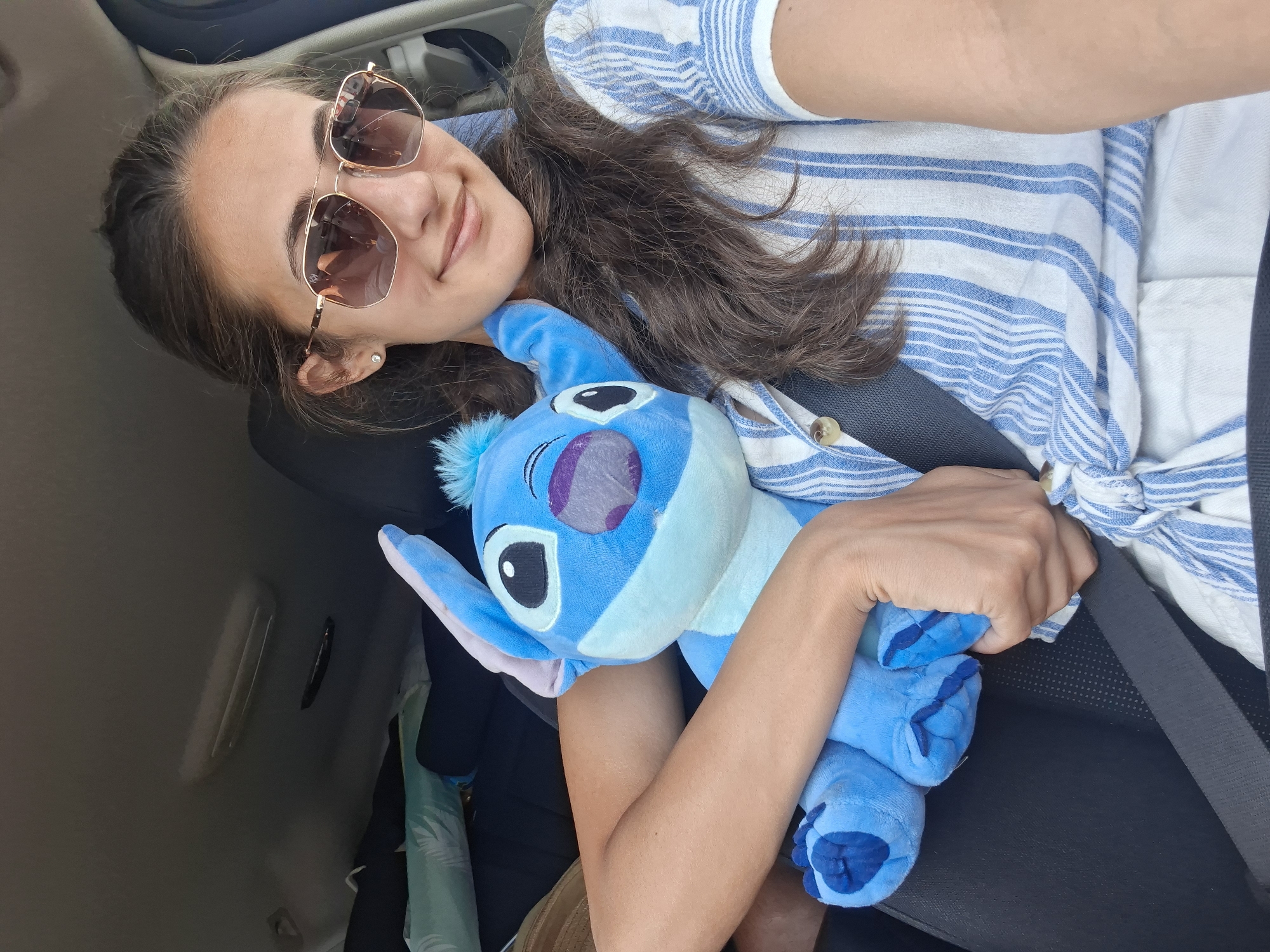 A young woman is sitting in the passenger seat of a car, holding a blue Stitch plush toy. She is wearing sunglasses and a blue and white striped shirt. The car appears to be packed with luggage. The woman is smiling at the camera, and the plush toy is positioned on her lap. The background shows the interior of the car, including the dashboard and the ceiling. The scene is brightly lit, suggesting it is daytime.