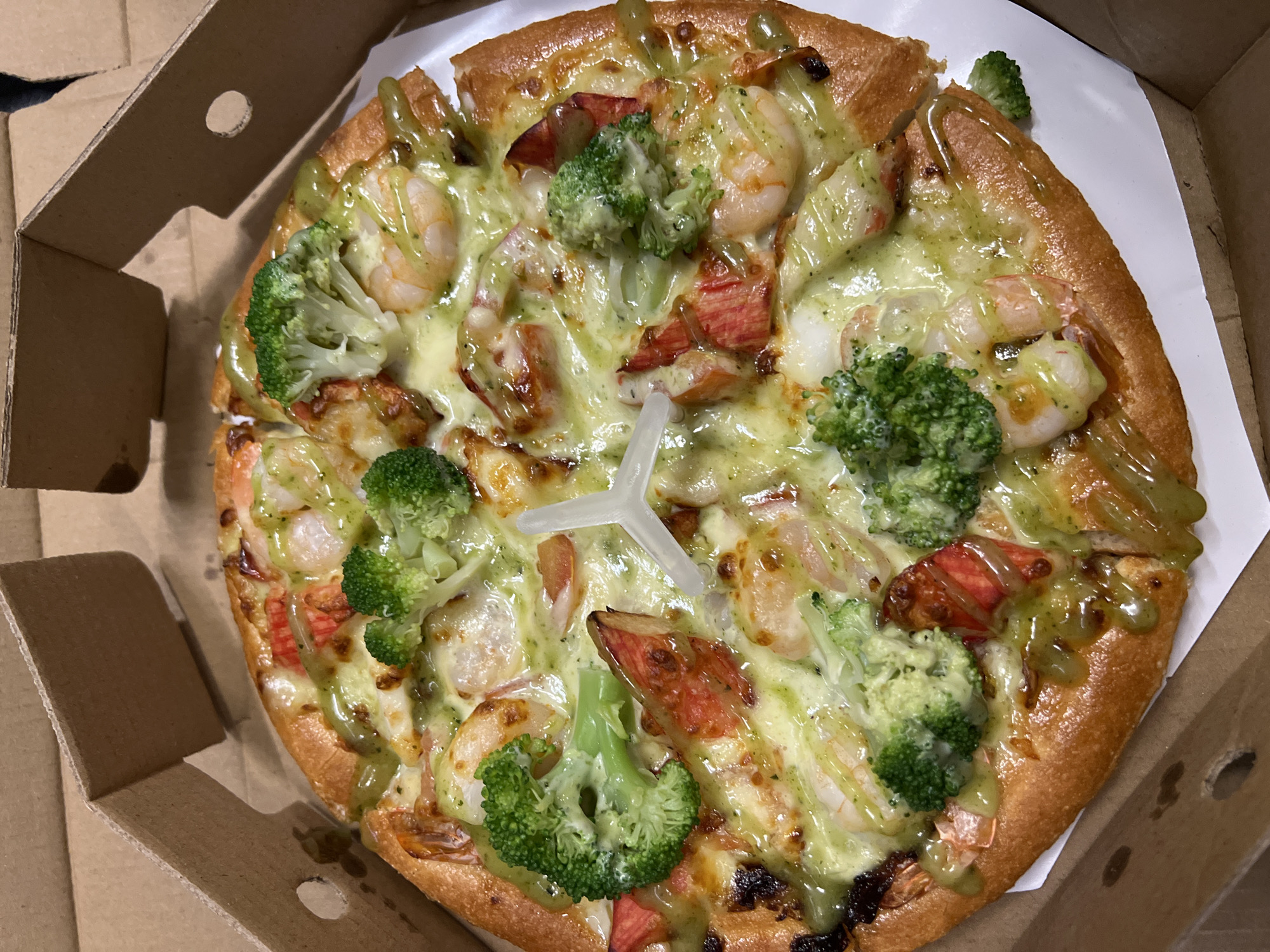 A close-up, overhead shot reveals a pizza inside a cardboard box. The pizza has a golden-brown crust and is topped with melted cheese, shrimp, broccoli florets, and what appears to be pieces of imitation crab meat. A drizzle of green sauce is also present on the pizza. A white plastic pizza saver is positioned in the center of the pizza. The pizza is likely in a take-out box, with the cardboard sides of the box visible around the pizza. There is no immediate indication of the image being located in Đà Nẵng, Vietnam, as there are no landmarks or other identifying characteristics to identify the location.