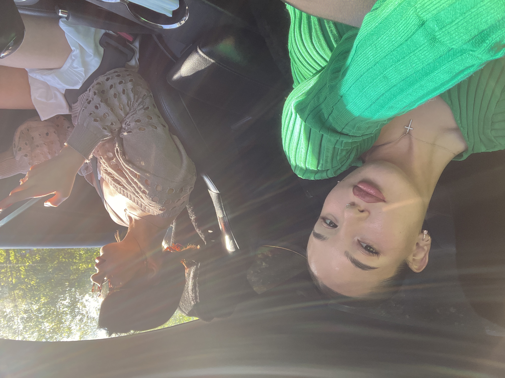 Two individuals are inside a vehicle. One wears a green ribbed top and a silver necklace with a cross. The other individual is partially visible, wearing a beige top with eyelet detailing. The setting appears to be during the day with bright sunlight, possibly in a suburban area given the trees visible through the car window. The location details of Perm, Russia cannot be confirmed from this image.