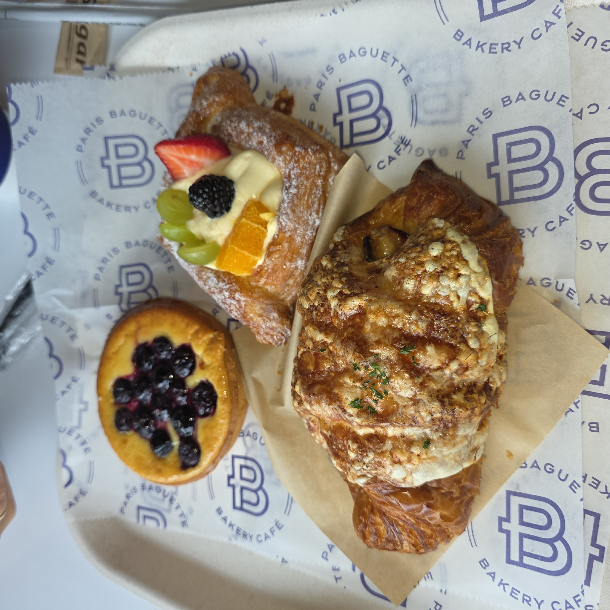 A tray of pastries sits on a white surface, likely a table. The pastries are arranged on a white paper liner with a repeating blue logo featuring a stylized 'b'.
The three pastries visible are:
1. A round tart topped with blueberries and a creamy filling.
2. A square-shaped pastry with a creamy filling, topped with sliced strawberries, green grapes, and a blackberry, dusted with powdered sugar.
3. A large, golden-brown croissant, possibly savory, with melted cheese and herbs on top.
A small brown packet, likely containing sugar, is visible in the upper right corner of the image. The overall lighting suggests an indoor setting. The image does not provide any information about a specific city or location within Beaverton, United States of America.