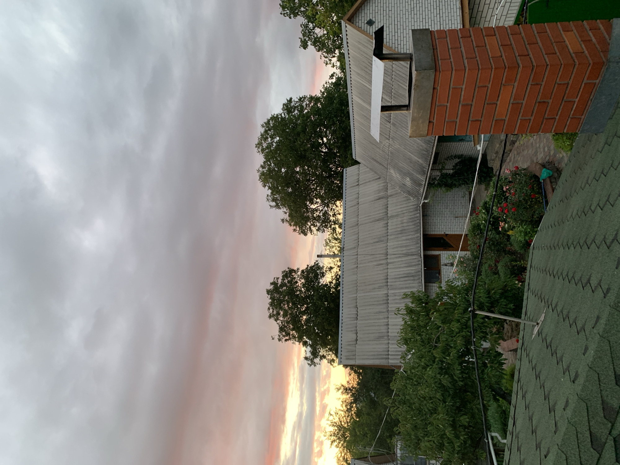 This image captures a rural scene in Tkhorivka, Ukraine, during a cloudy sunset. The sky is filled with dramatic, overcast clouds tinged with pink and yellow hues from the setting sun, creating a soft, diffused light. In the foreground, a green asphalt shingle roof is visible, with a metallic object attached to it, possibly an antenna base. To the right, a prominent brick chimney stands tall, made of reddish-orange bricks and topped with a metallic cap.
Behind the chimney, a large, gray slate-roofed house is partially visible, with white brick walls and wooden window frames. To the left of this house, another similar structure with a gray slate roof can be seen. Several lush green trees with dense foliage dot the landscape, adding depth and natural elements to the scene. In the garden area between the houses, vibrant red roses are in bloom, accompanied by other greenery and plants, suggesting a well-maintained outdoor space. A pathway made of brick or stone winds through the garden. Power lines are also visible, stretching across the scene. There are no people present in the image.