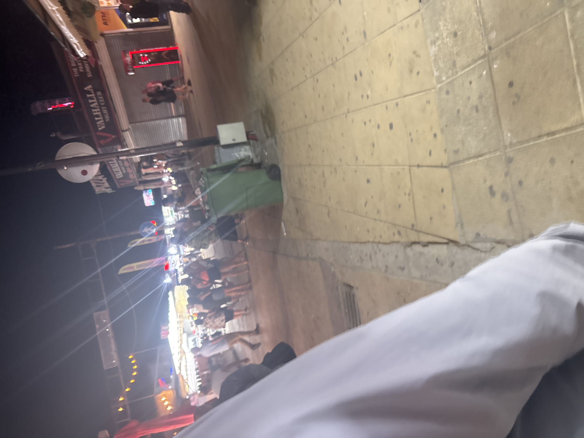This is a nighttime outdoor scene on a street in Nesebar, Bulgaria. The image is slightly blurred. In the background, there are several establishments with illuminated signs. One sign reads "VALHALLA NIGHT CLUB" with a stylized "V" logo. Adjacent to this, another sign is partially visible with text that appears to be "THE BIGGEST PARTY SUNNY BEACH". To the left of Valhalla, there is a sign that says "PIZZA".
In front of these establishments, a group of people are visible. Some are standing and talking, while others appear to be walking. They are dressed in casual clothing. There is a green garbage bin with wheels on the left side of the foreground. The ground is paved with square tiles, some of which appear to be wet or stained. The overall impression is a lively street scene at night, likely a tourist or entertainment area.