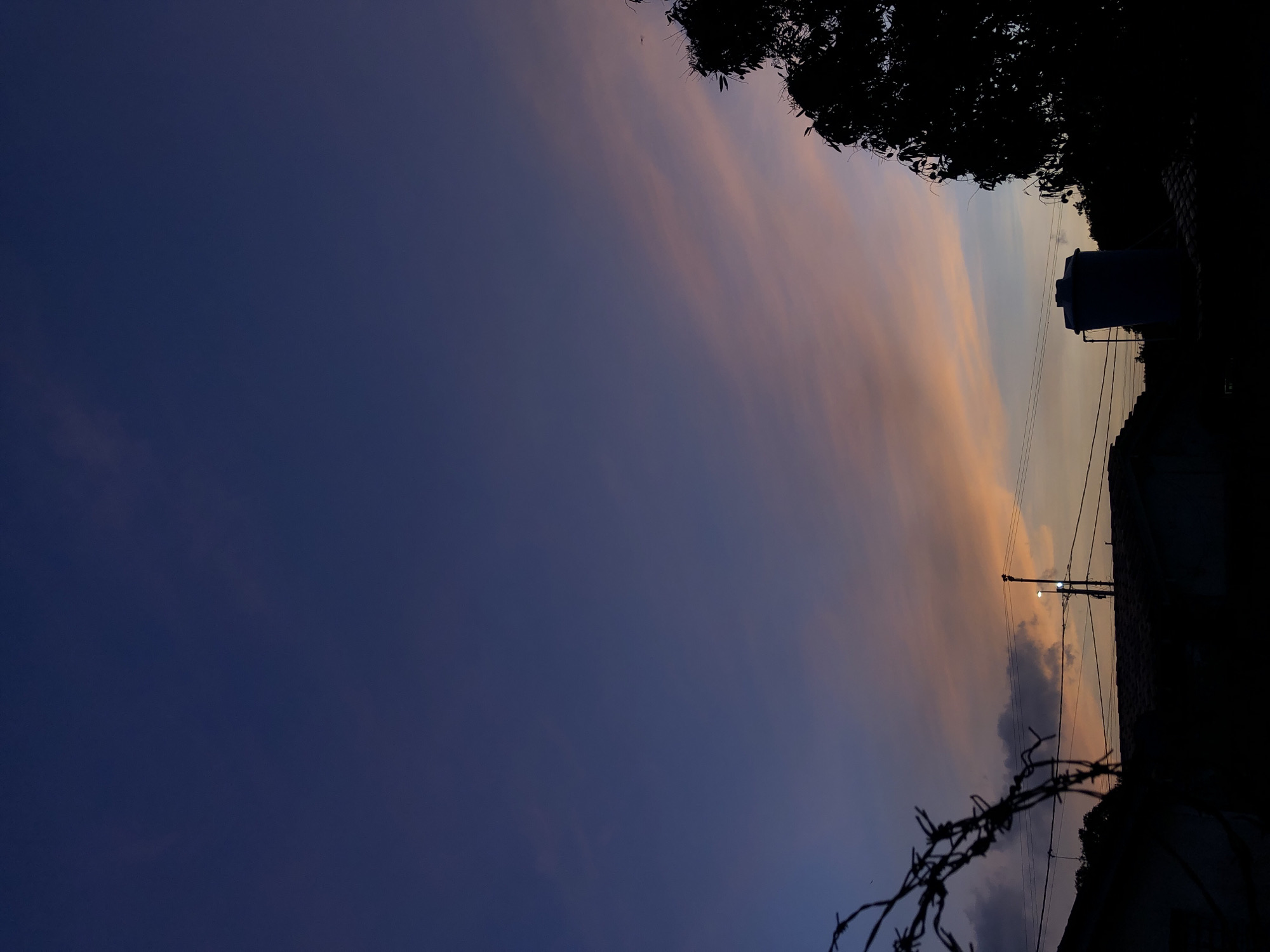 The image presents a twilight scene, possibly in Altagracia, Venezuela, though specific location verification is impossible without further information. The sky transitions from a deep blue to a lighter, orange-tinted gradient near the horizon. Silhouetted against the sky are various elements: a large tree branch at the top right, power lines stretching across the frame, a water tank atop a building, a streetlight, and a section of wall or fence with barbed wire. The lower right corner is predominantly dark, obscuring details on the ground level. There are no visible people or interactions depicted. The setting evokes a quiet, possibly rural or suburban neighborhood environment at dusk.