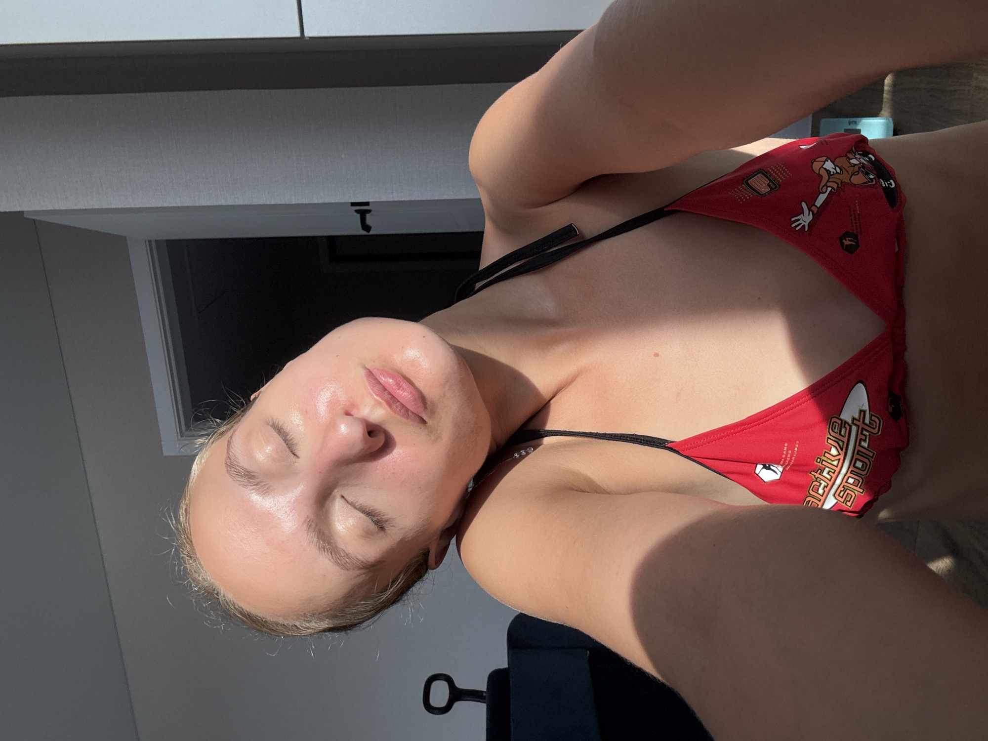 A young woman with her eyes closed is bathed in warm sunlight. She is wearing a red bikini top with white and yellow lettering and cartoon graphics. Her head is tilted back, and her skin appears to be glowing from the sun. The background is dimly lit, with a doorway visible on the right side of the image. There is a shadow cast across the woman from the left, suggesting the sun is positioned at an angle. The overall scene appears to be indoors, possibly in a bedroom or living area. The city of Подшивалово, Russia, is not depicted or suggested in this image.