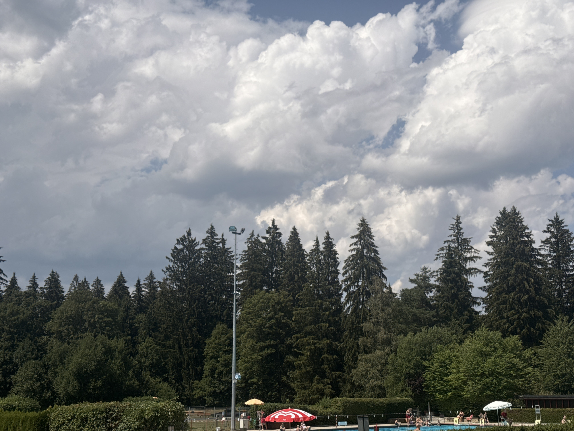 The image depicts an outdoor swimming pool area on a cloudy day. In the foreground, a blue swimming pool is visible with several people in and around it. There are red and white striped umbrellas providing shade, and some people are seated under them. Some individuals are swimming or wading in the pool. To the left of the pool, a tall lamppost stands with a clock and what appears to be a camera or sensor attached to it. Behind the pool area, a dense line of tall evergreen trees forms a backdrop, with some deciduous trees interspersed. The sky above is filled with dramatic, fluffy white clouds against a pale blue sky. The overall scene suggests a recreational area surrounded by nature, likely located in a temperate climate. Based on the context of the query, this scene is likely the Piscine de la Blanchisserie in Le Locle, Switzerland, a well-known outdoor swimming facility.