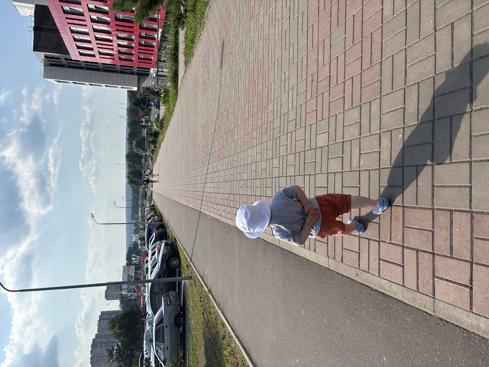 A young child wearing a white bucket hat, gray t-shirt, orange shorts, and blue clogs walks away from the camera on a paved sidewalk. The sidewalk is made of interlocking rectangular bricks, arranged in a herringbone pattern with alternating red and gray colors. The child's hands are clasped behind their back. To the left of the sidewalk is an asphalt road and a parking lot filled with various cars, including sedans and SUVs. Beyond the parking lot, there are tall, modern apartment buildings, some with red accents, and a line of trees. In the background, a clear blue sky with scattered white clouds is visible. The scene appears to be in a residential or mixed-use area of Naberezhnye Chelny, Russia, given the presence of distinctive architecture and Russian car models. The lighting suggests it is a sunny day.