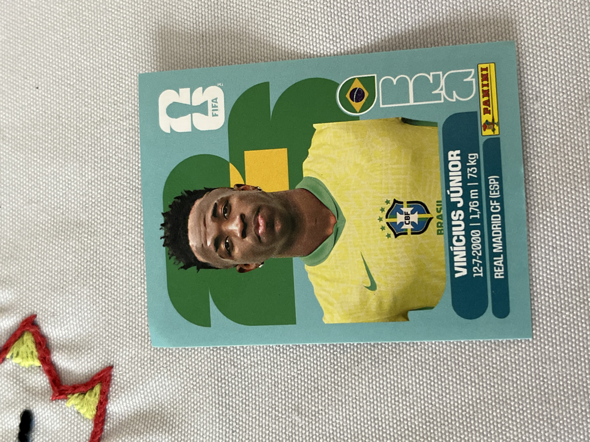 The image shows a Vinícius Júnior Panini FIFA trading card. The card features a photo of Vinícius Júnior wearing a yellow Brazil national team jersey with the CBF logo. Text on the card indicates his name, date of birth (12-7-2000), height (1.76 m), weight (73 kg), and club (Real Madrid CF, ESP). The card also displays the FIFA logo, the Panini logo, and other design elements. The card is photographed on a textured surface, with the edge of a craft project with red and yellow stitching visible in the lower left corner.