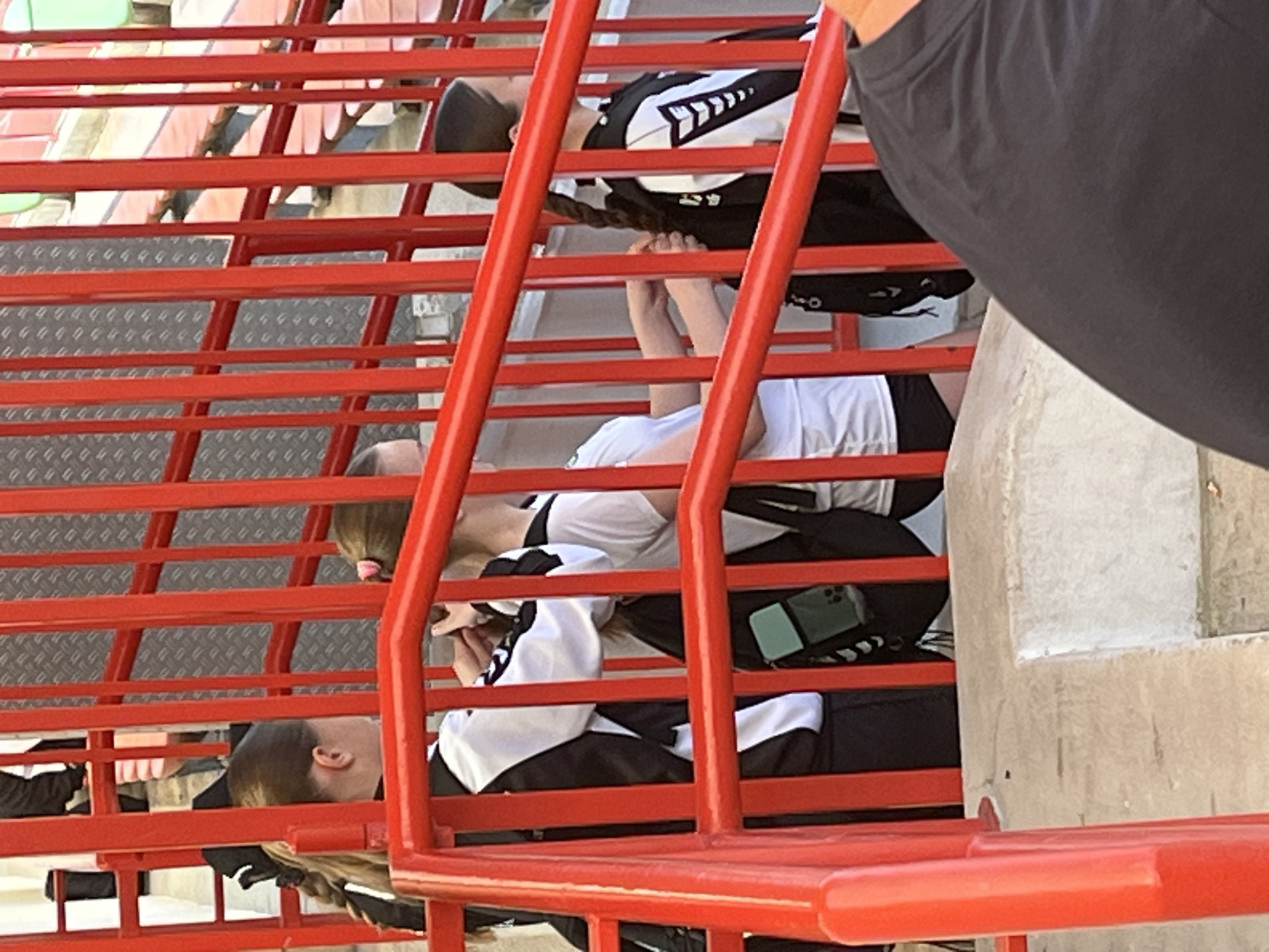 Three people are on a red metal staircase in Kelsterbach, Germany. Two people wear black and white jackets and the third wears a white shirt and black shorts. One of the jacket-wearers is braiding the hair of another. A cell phone rests on one of the steps. A person wearing a dark grey shirt stands beside the staircase. The environment includes a metal staircase and a light-colored concrete support.
