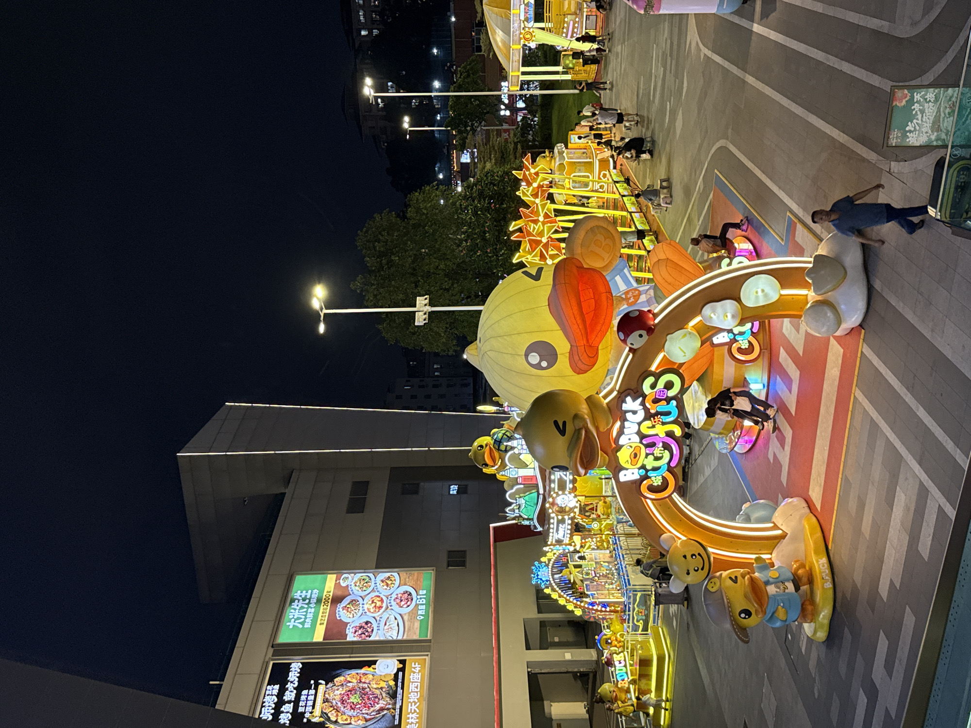 This is a night-time, wide-angle shot of an outdoor plaza in Tianhe District, China, likely a shopping or entertainment area. The scene is brightly lit with decorative lights and illuminated signs, suggesting a festive atmosphere, possibly a holiday or event.
The primary focus is a large, colorful, cartoonish duck-themed decoration that curves around the foreground. This decoration features large inflatable ducks, smaller duck figures, and a bright, neon-like sign that reads "BDuck Cityfirs". Several people are visible in the background, some walking through the plaza and others gathered around amusement rides or stalls, indicating a public gathering. The ground is paved with light and dark grey tiles, marked with white lines, resembling a road or walkway.
To the left of the duck decoration, a building with a modern architectural design is visible, with lights running along its edges. Below the building, there are illuminated billboards with Chinese text and images of food, suggesting restaurants or shops. One billboard prominently displays "大米先生" (Mr. Rice) and advertises dishes, and another seems to be for a food establishment with a prominent image of a hot pot.
The overall impression is a vibrant and lively urban setting at night, with a focus on entertainment and consumer activities. The lighting is a key element, creating a cheerful and inviting ambiance. The weather appears clear, given the lack of visible precipitation and the presence of people outdoors.