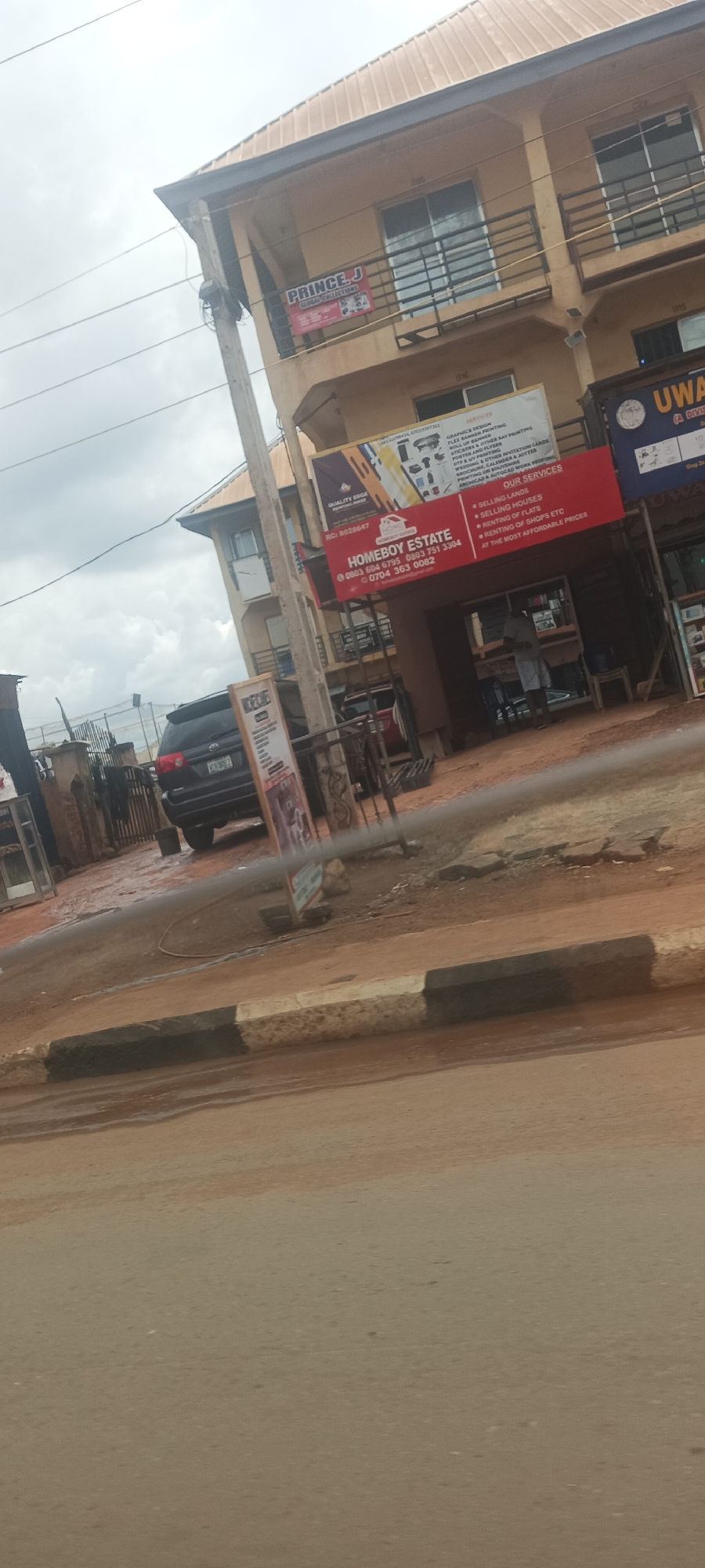 The image depicts a street scene in what appears to be a commercial area in Enugu, Nigeria. It seems to have rained recently, as the road and surrounding ground are wet. In the background, there is a black SUV parked near a fence. Several businesses are visible, indicated by signs: "PRINCE. J GLOBAL COLLECTIONS" on the upper floor and "HOMEBOY ESTATE" and "UWA (A DIVISION)" on the ground floor. There are power lines and a utility pole. A person is vaguely visible within the entrance of the HOMEBOY ESTATE building. There is a sidewalk with black and white painted curbs along the road.