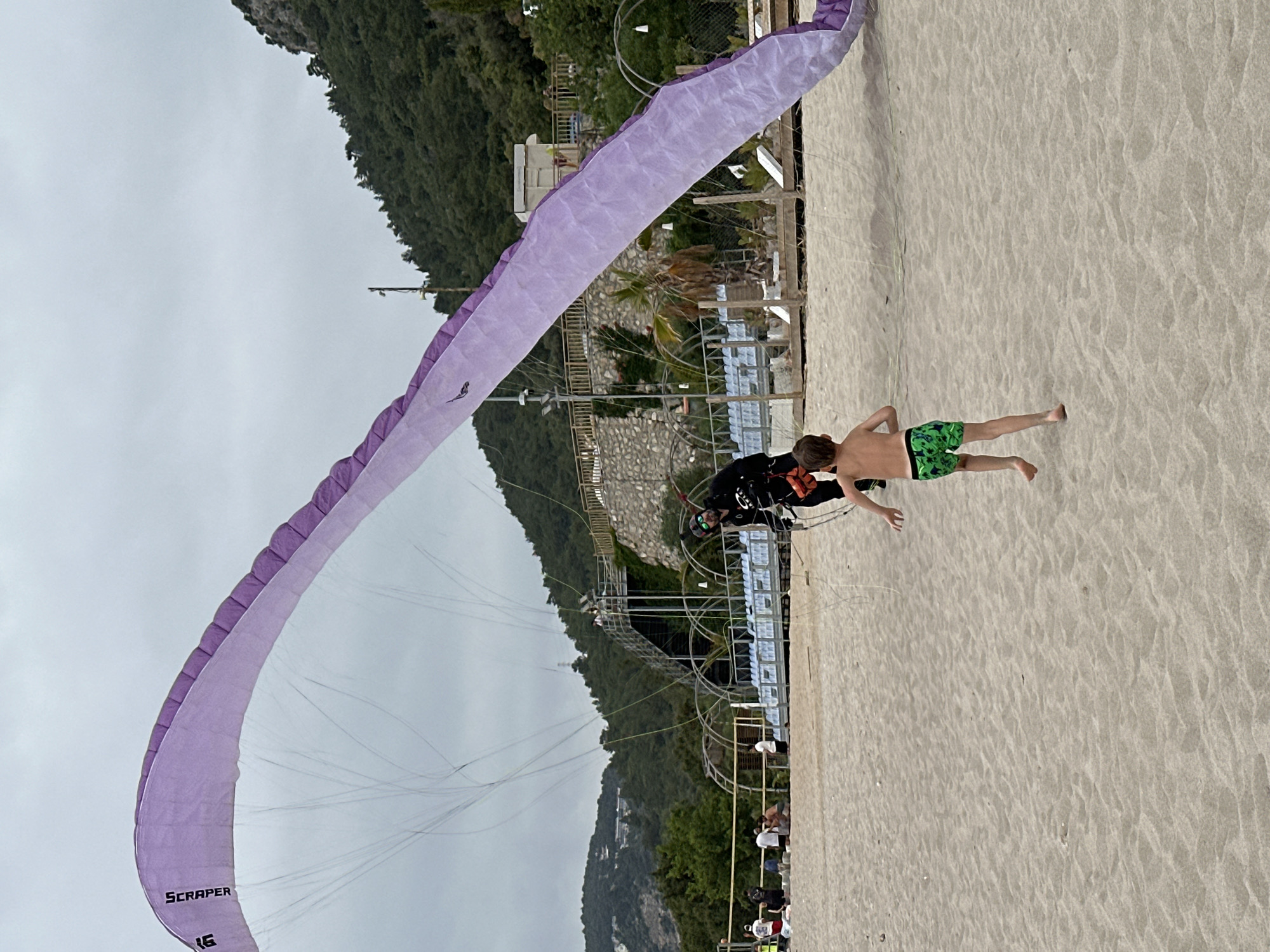 On a cloudy day in Fethiye, Turkey, a paraglider is taking off from a beach. A young boy in green and black swim trunks runs along the sand. He is positioned between the paraglider and its pilot, who is harnessed up and wearing a helmet and sunglasses. The paraglider is a light purple color with the word "SCRAPER" printed on the edge. The beach is a light tan color, and in the background, there is a structure that appears to be an observation point with a white railing. Behind the structure, a steep, green, tree-covered hill rises up. Further in the background is the cloudy sky. Other people can be seen in the distance on the beach near the structure.