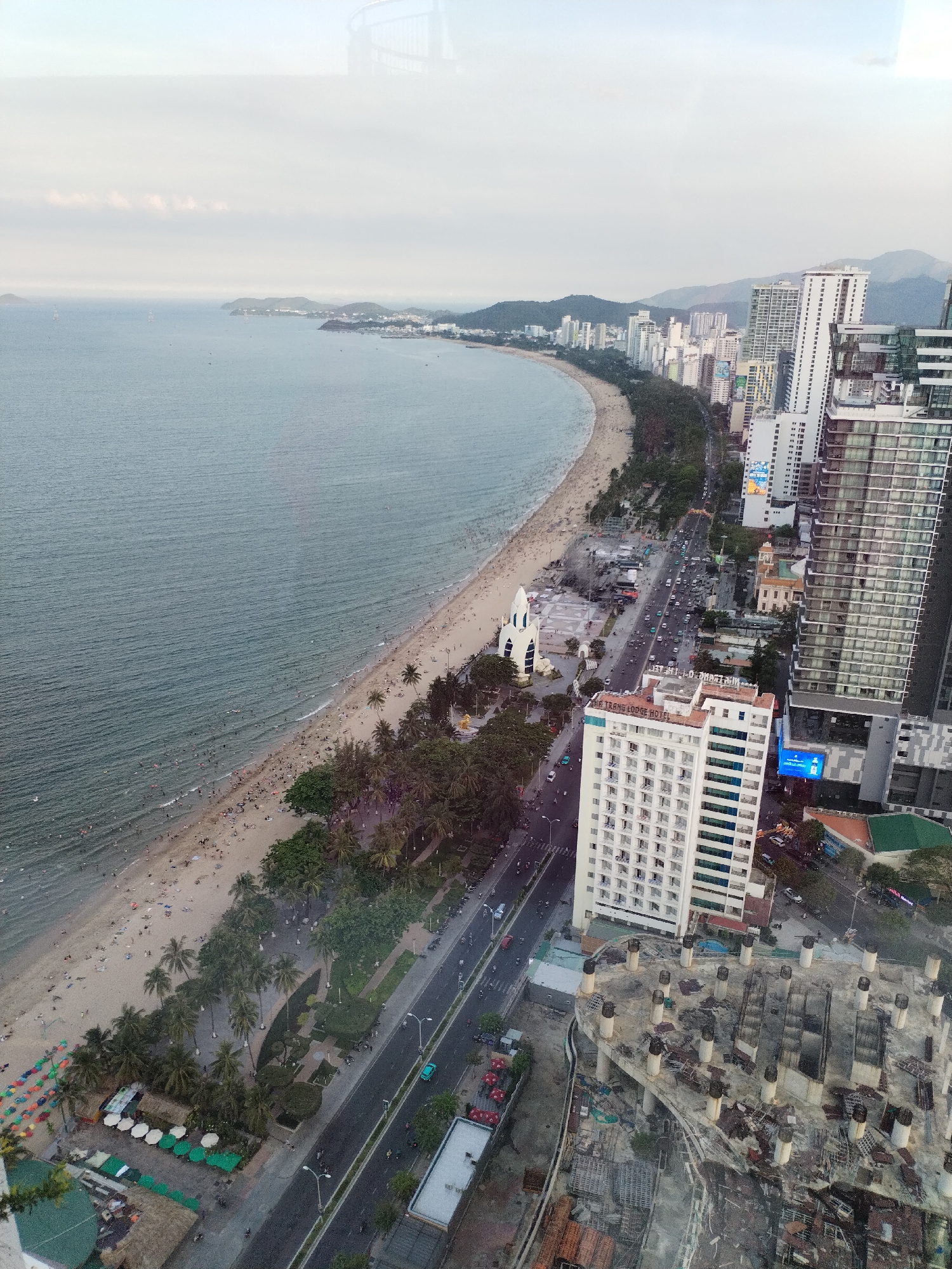 This is an aerial view of the Nha Trang coastline in Vietnam. A wide, sandy beach is visible, populated by numerous beachgoers. The sea, a calm expanse of muted blue, gently laps the shore. Along the beach, a strip of palm trees provides shade. Parallel to the beach, a multi-lane road carries a moderate flow of traffic, including cars, motorcycles, and buses. Numerous high-rise buildings, characteristic of a coastal city, line the road and extend inland. One prominent building, "The Tran Lodge Hotel," is clearly labeled. In the foreground, an unfinished concrete structure with exposed rebar and cylindrical supports suggests ongoing construction. The overall scene depicts a vibrant urban coastal environment with recreational activities and development.
